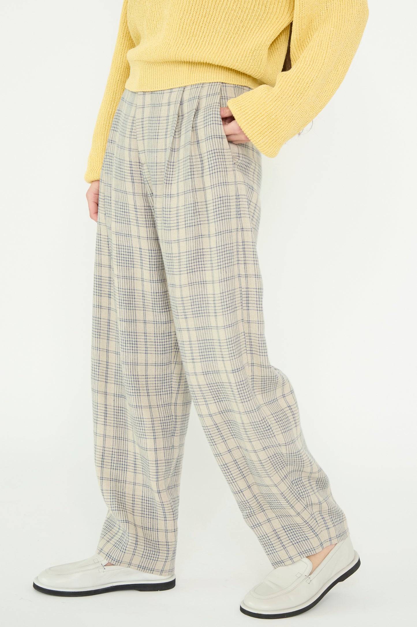 A model wears a yellow sweater, Cristaseya's Linen Double Pleated Wide Pant in Cream with Petrol Checks, and white shoes while standing against a plain background.