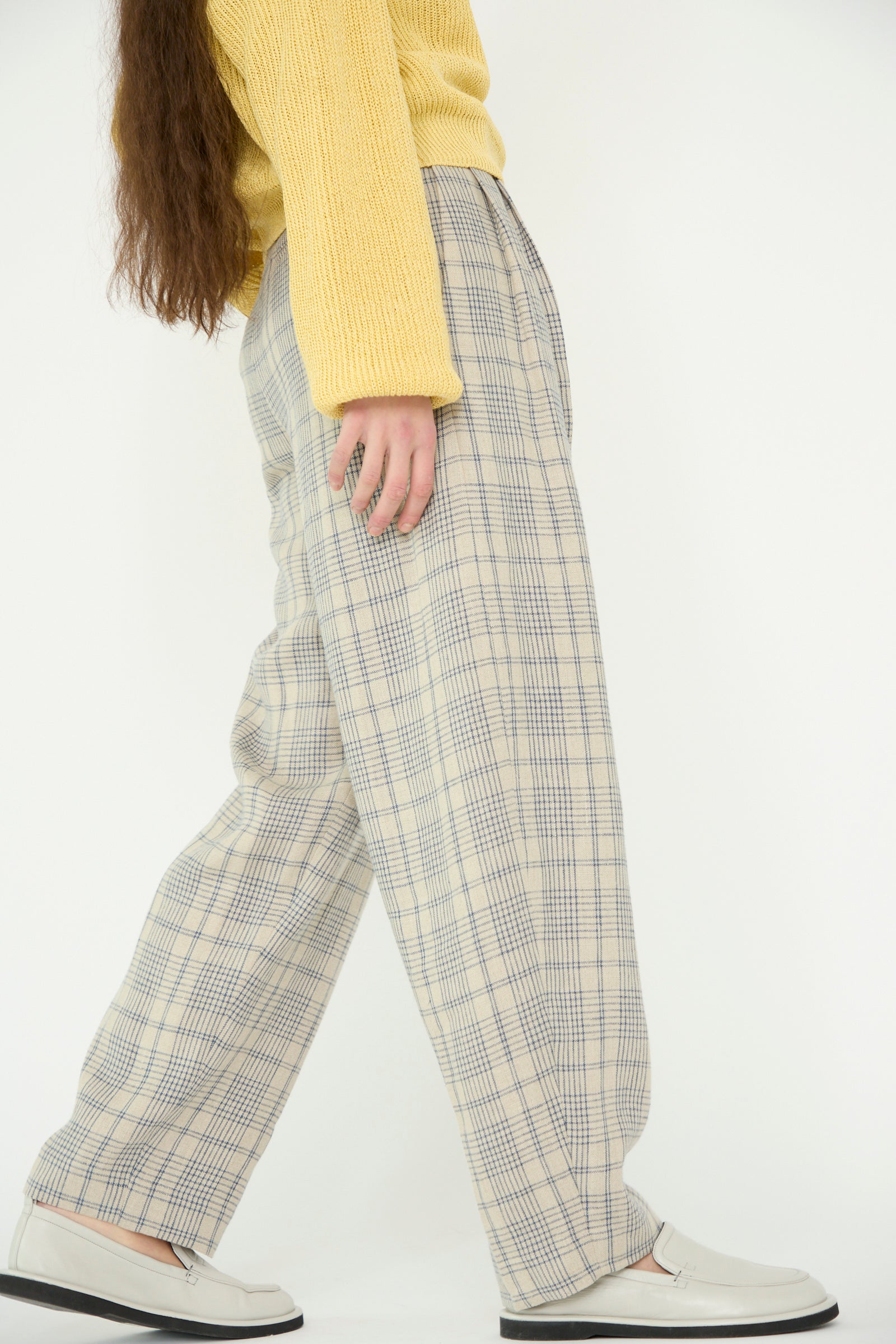 A person in a yellow sweater and white loafers steps to the right, showcasing Cristaseya's Linen Double Pleated Wide Pant in Cream with Petrol Checks against a white backdrop. 