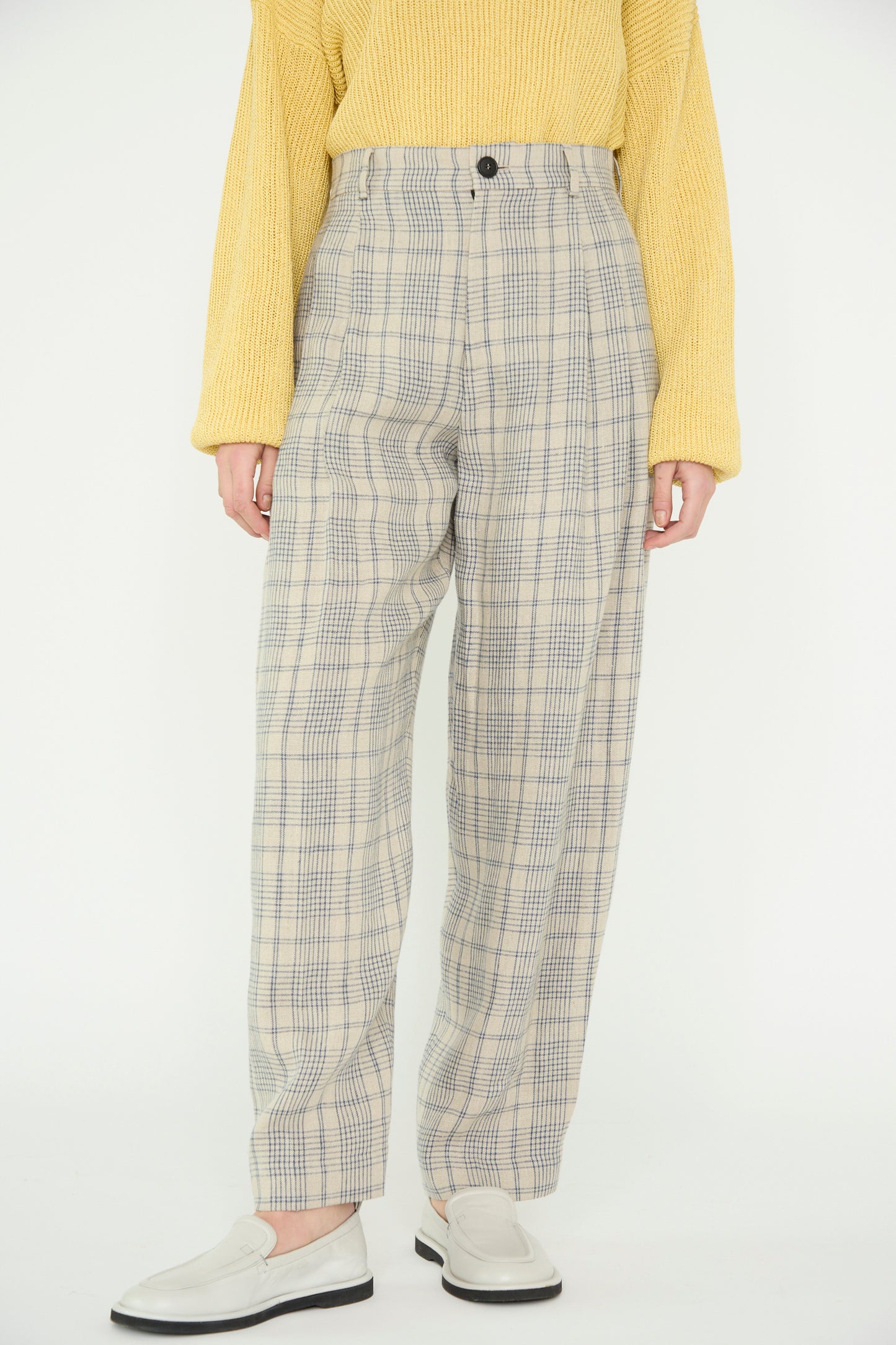 A model in Cristaseya's Linen Double Pleated Wide Pant in Cream with Petrol Checks and a yellow sweater, stands against a plain backdrop, with white shoes. 