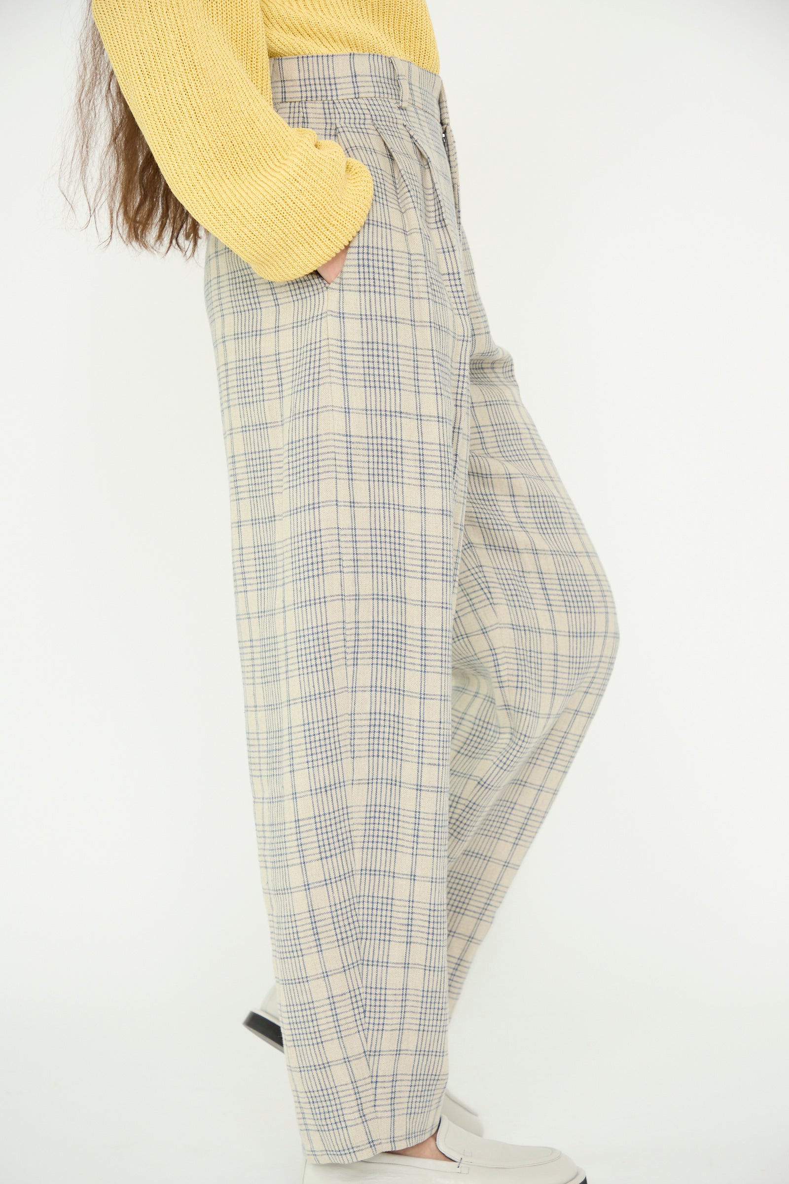 A model wearing a yellow sweater and Cristaseya's Linen Double Pleated Wide Pant in Cream with Petrol Checks, with hands in pockets against a plain backdrop. 