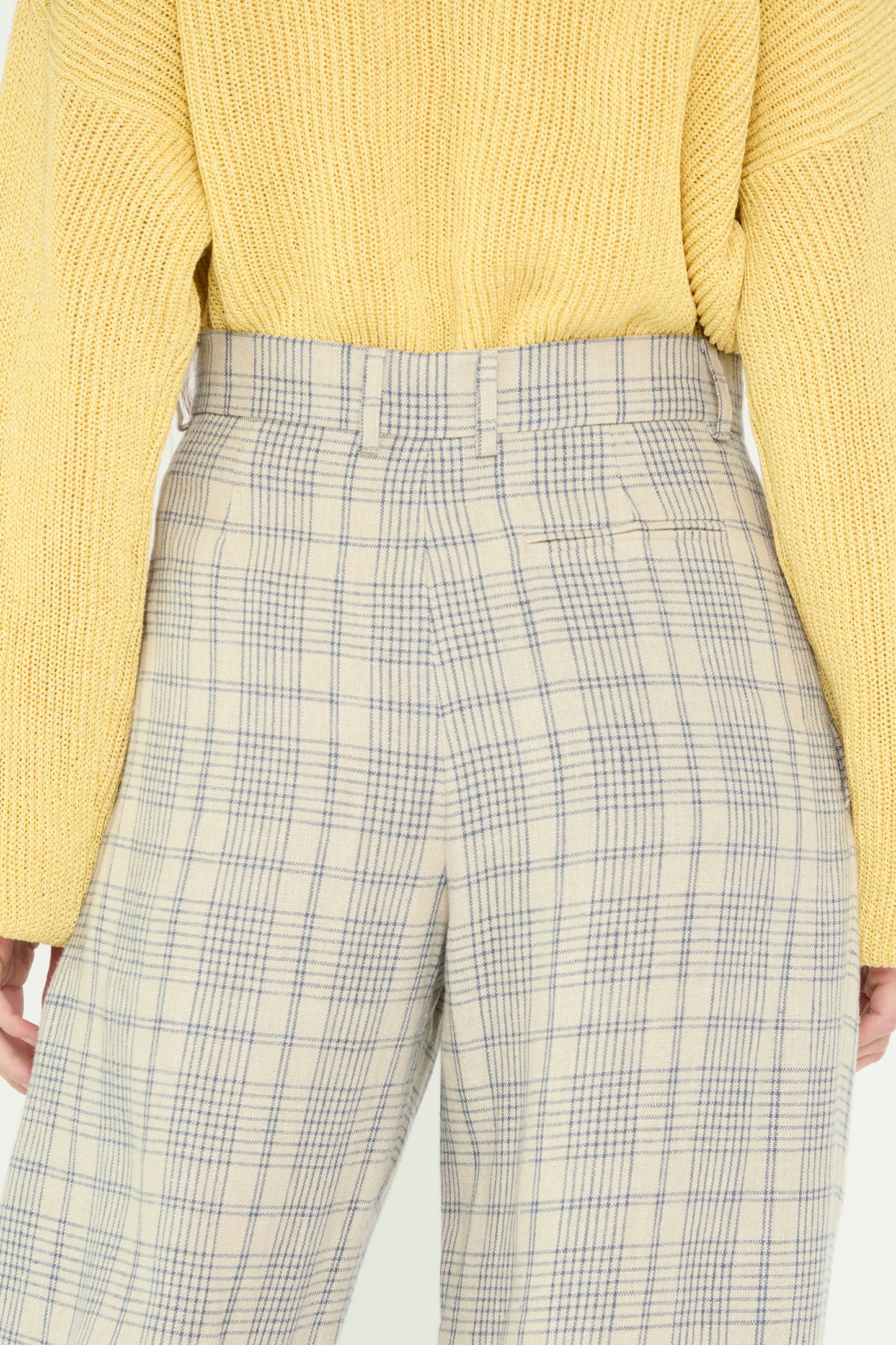 A close-up of a model wearing a yellow sweater and Cristaseya's Linen Double Pleated Wide Pant in Cream with Petrol Checks stands with their back to the camera.