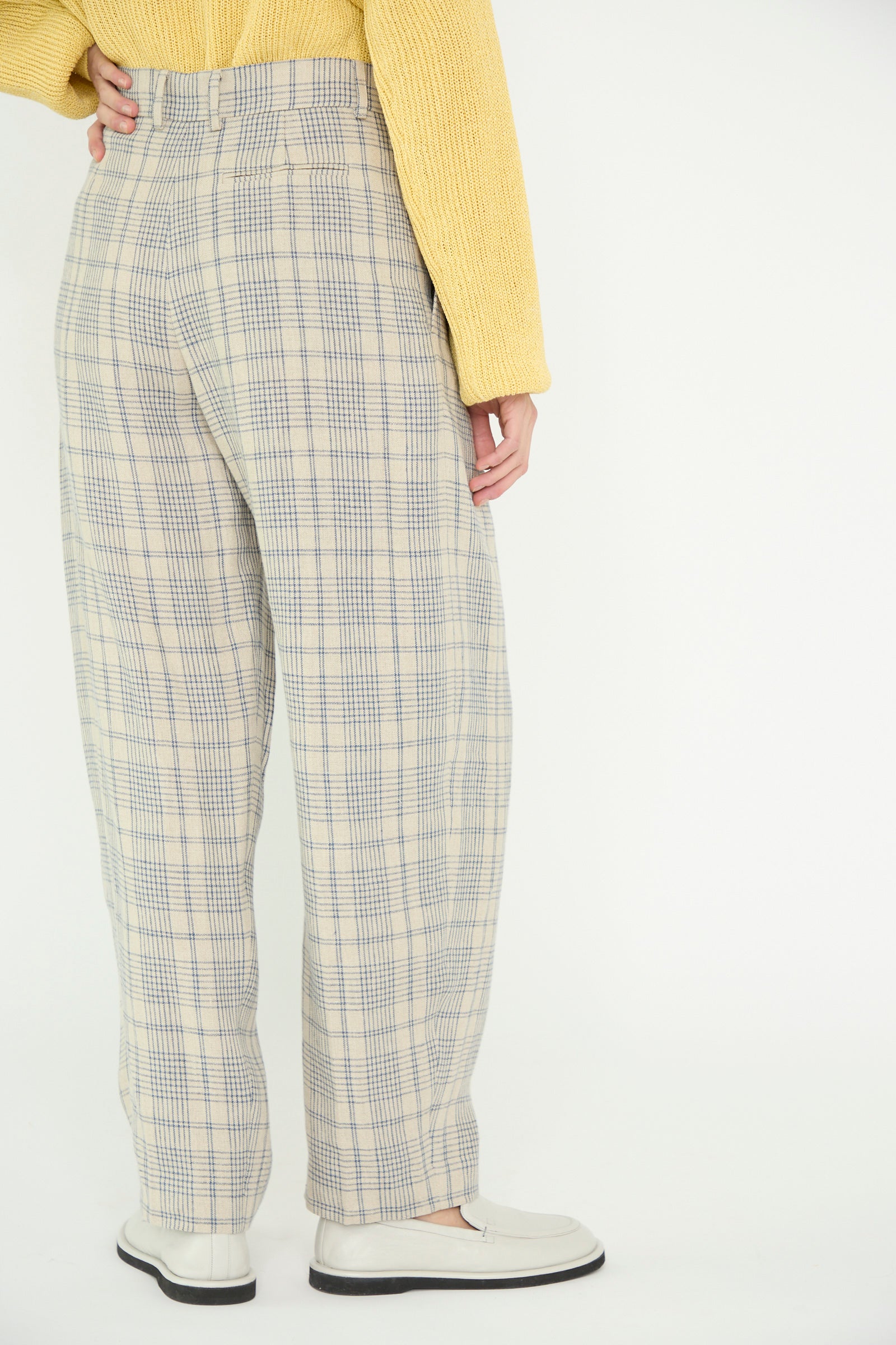 Against a plain background, a model facing back wearing a yellow sweater with Cristaseya's Linen Double Pleated Wide Pant in Cream with Petrol Checks and white shoes.