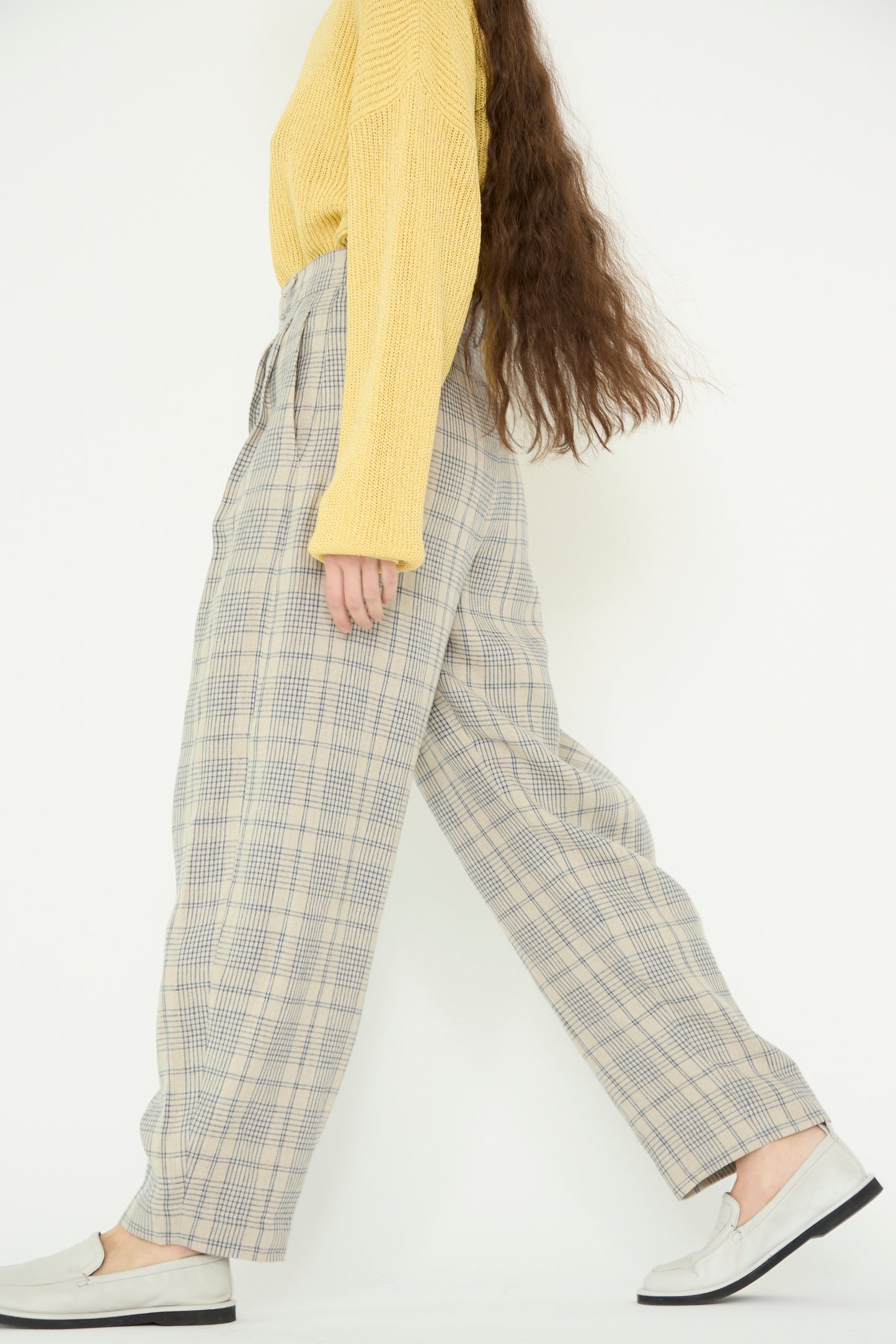 A model in a yellow sweater and Cristaseya linen double-pleated wide pants with petrol checks walks to the right against a white backdrop. 