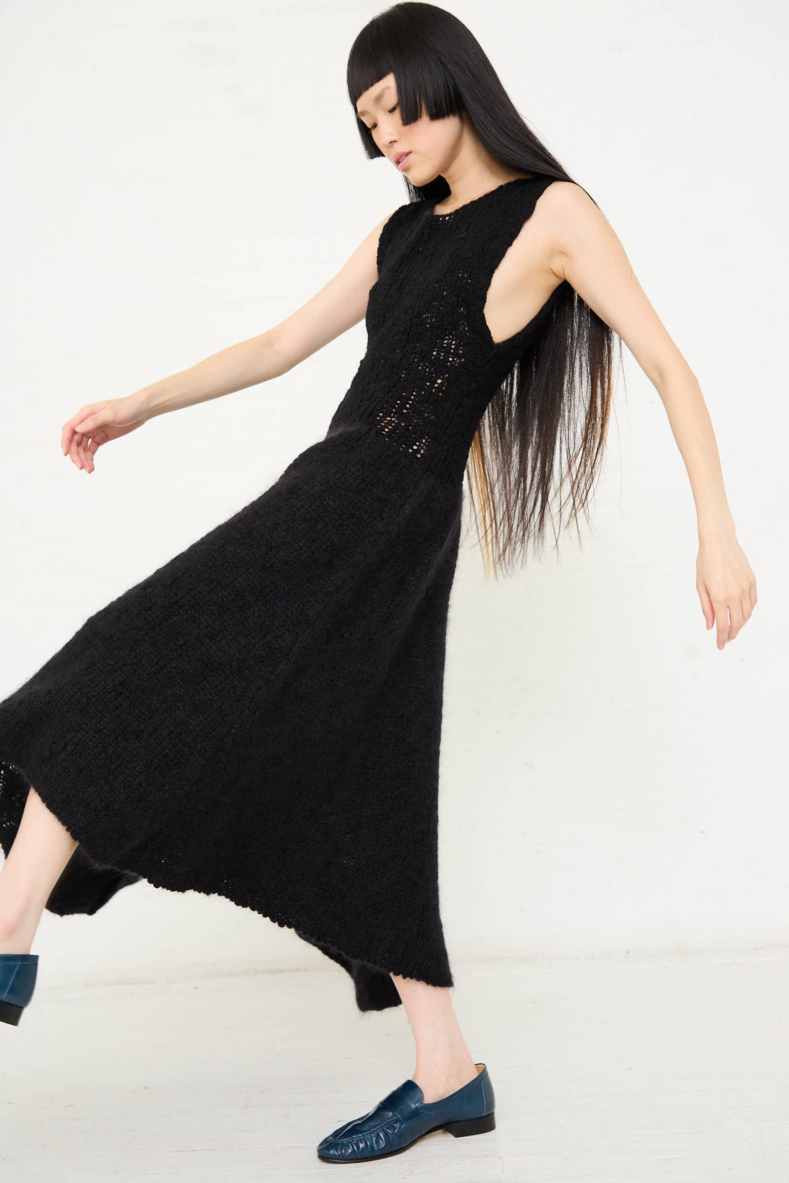 A person with long black hair wearing the Cristaseya Merino and Mohair Asymmetrical Long Knit Dress in Black stands on a white floor against a plain white background, complemented by blue shoes.