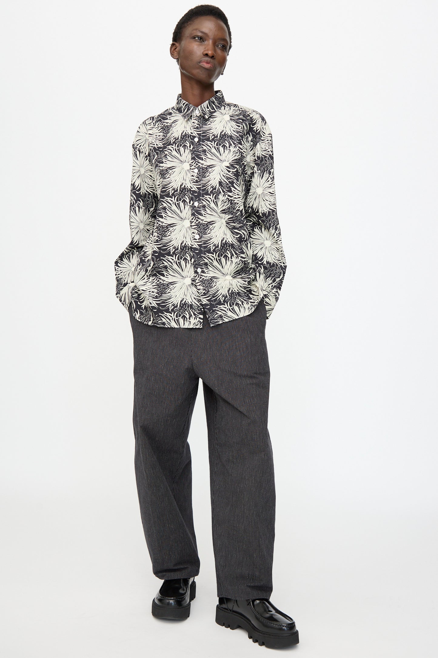 A person stands against a white background, wearing a black and white floral shirt, Cristaseya Moroccan Pyjama Pants in Black with White Stripes, and black chunky shoes.