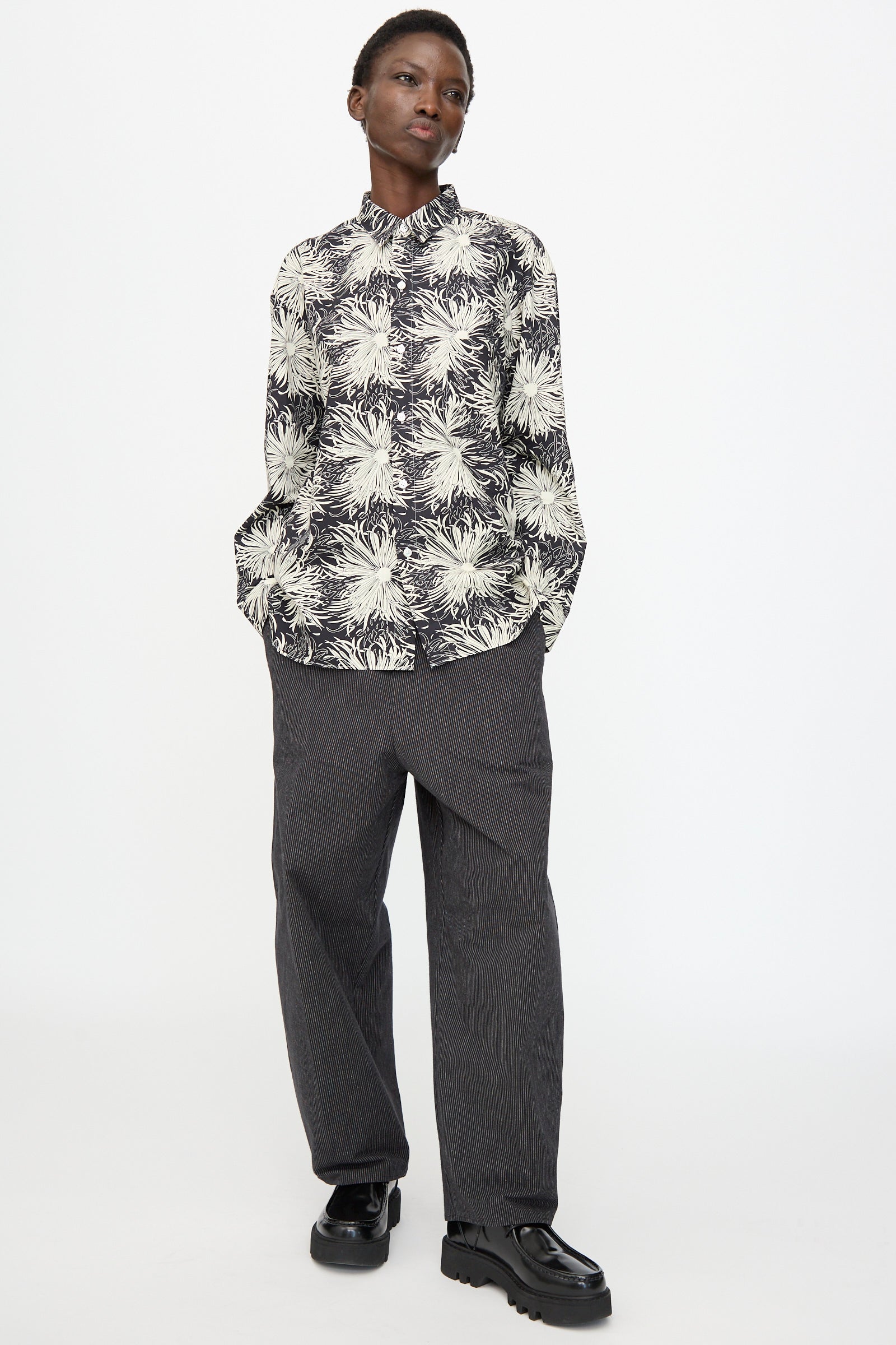 A person stands against a white background, wearing a black and white floral shirt, Cristaseya Moroccan Pyjama Pants in Black with White Stripes, and black chunky shoes.