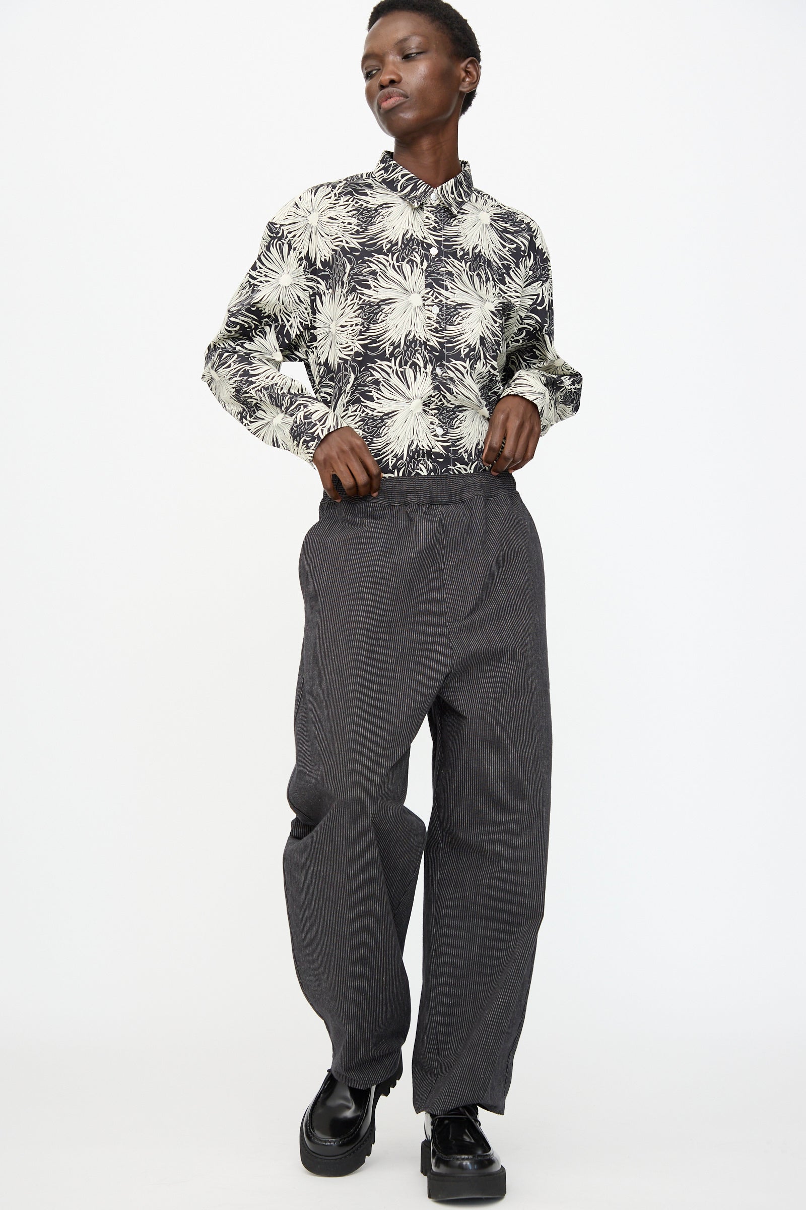 A person stands against a plain background wearing a black and white floral button-up shirt, Cristaseya Moroccan Pyjama Pants in Black with White Stripes, and black shoes.