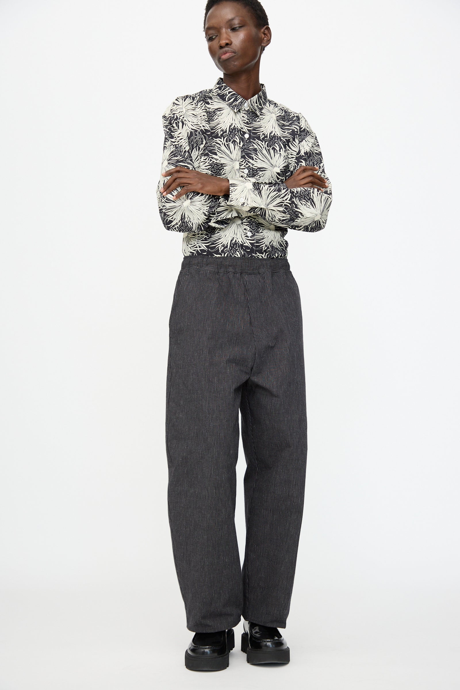 A person stands with arms crossed wearing Cristaseya Moroccan Pyjama Pants in Black with White Stripes, a black and white patterned long-sleeve shirt, and black shoes against a plain white background.