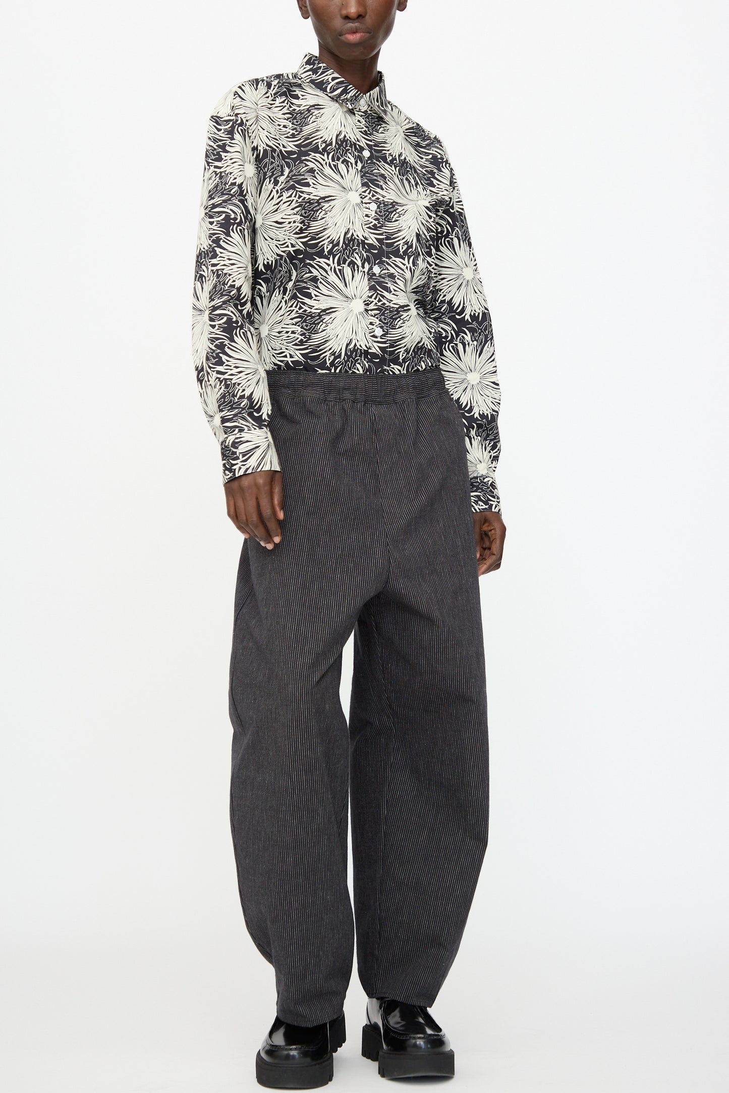 A person wearing Cristaseya's Moroccan Pyjama Pants in Black with White Stripes, a black and white floral button-up shirt, and black platform shoes stands against a plain white background.