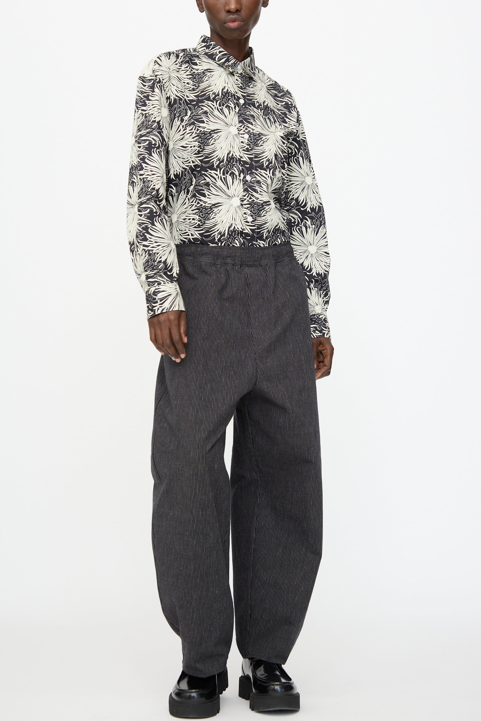 A person wearing Cristaseya's Moroccan Pyjama Pants in Black with White Stripes, a black and white floral button-up shirt, and black platform shoes stands against a plain white background.