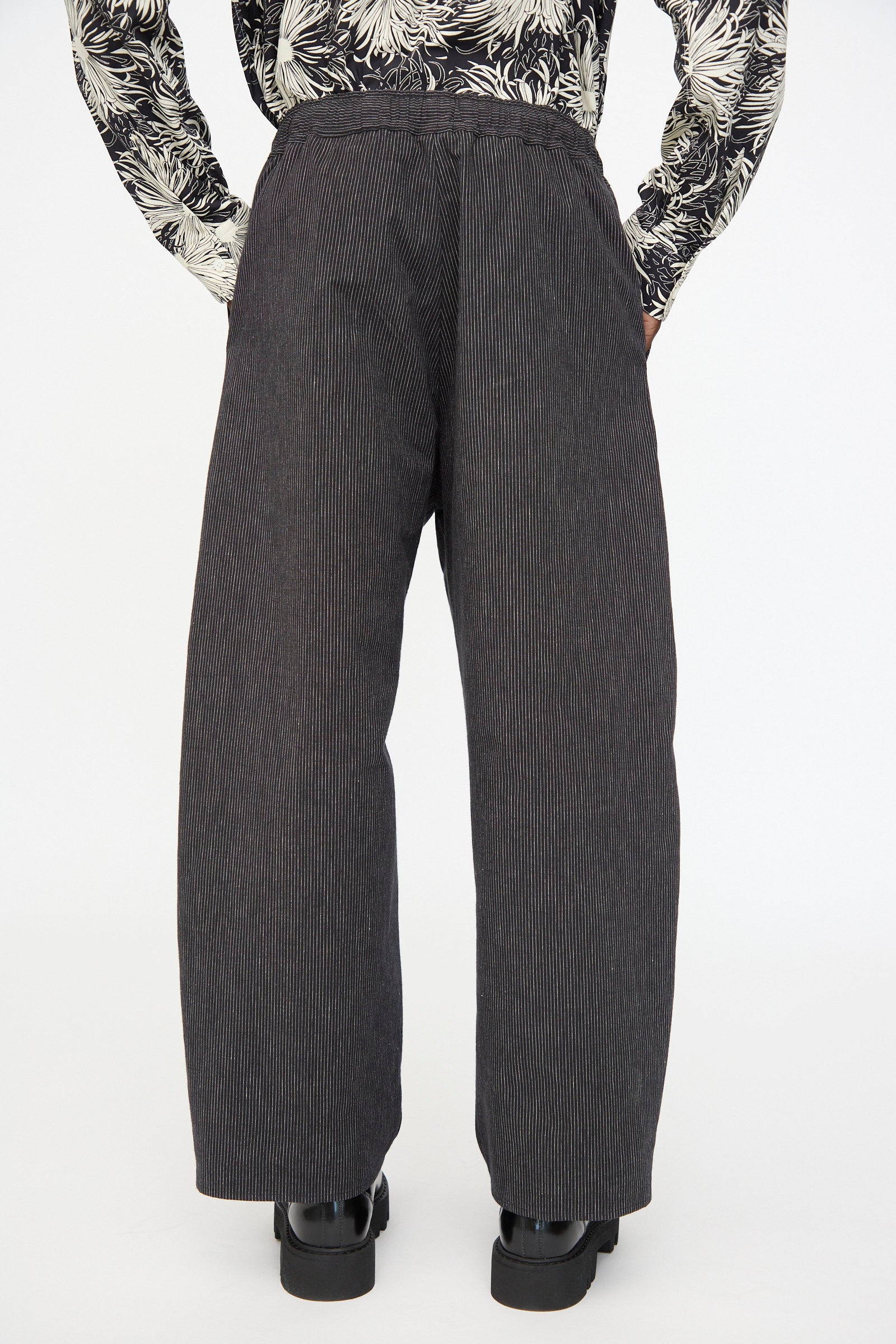 A person wearing Cristaseya Moroccan Pyjama Pants in Black with White Stripes, black shoes, and a patterned long-sleeve shirt, viewed from the back against a plain background.