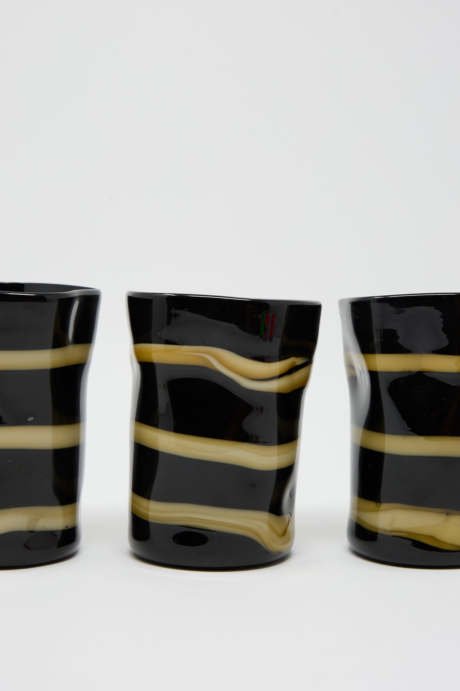 Three Cristaseya Murano Glass mugs in black with ocher stripes, featuring irregular wavy shapes, are lined up against a plain white background.