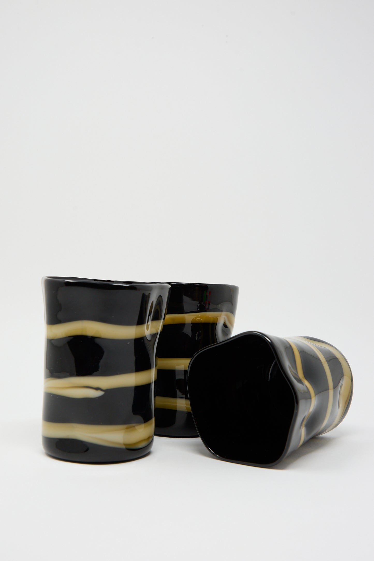 Three Murano Glass tumblers in black with ocher stripes by Cristaseya. Two stand upright, while the third is lying on its side.