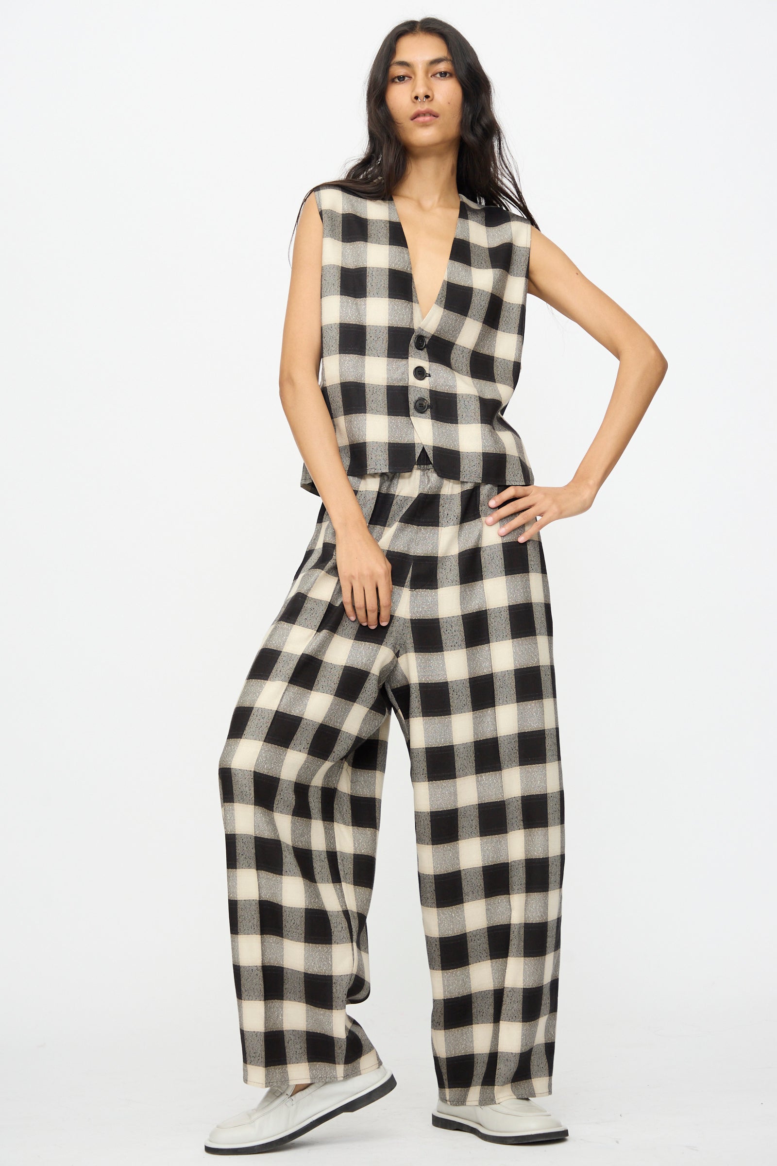 A person stands against a plain background wearing Cristaseya’s Oversized Pyjama Pants in Black and Cream Checks, paired with a matching sleeveless checkered bouclé vest and white shoes.