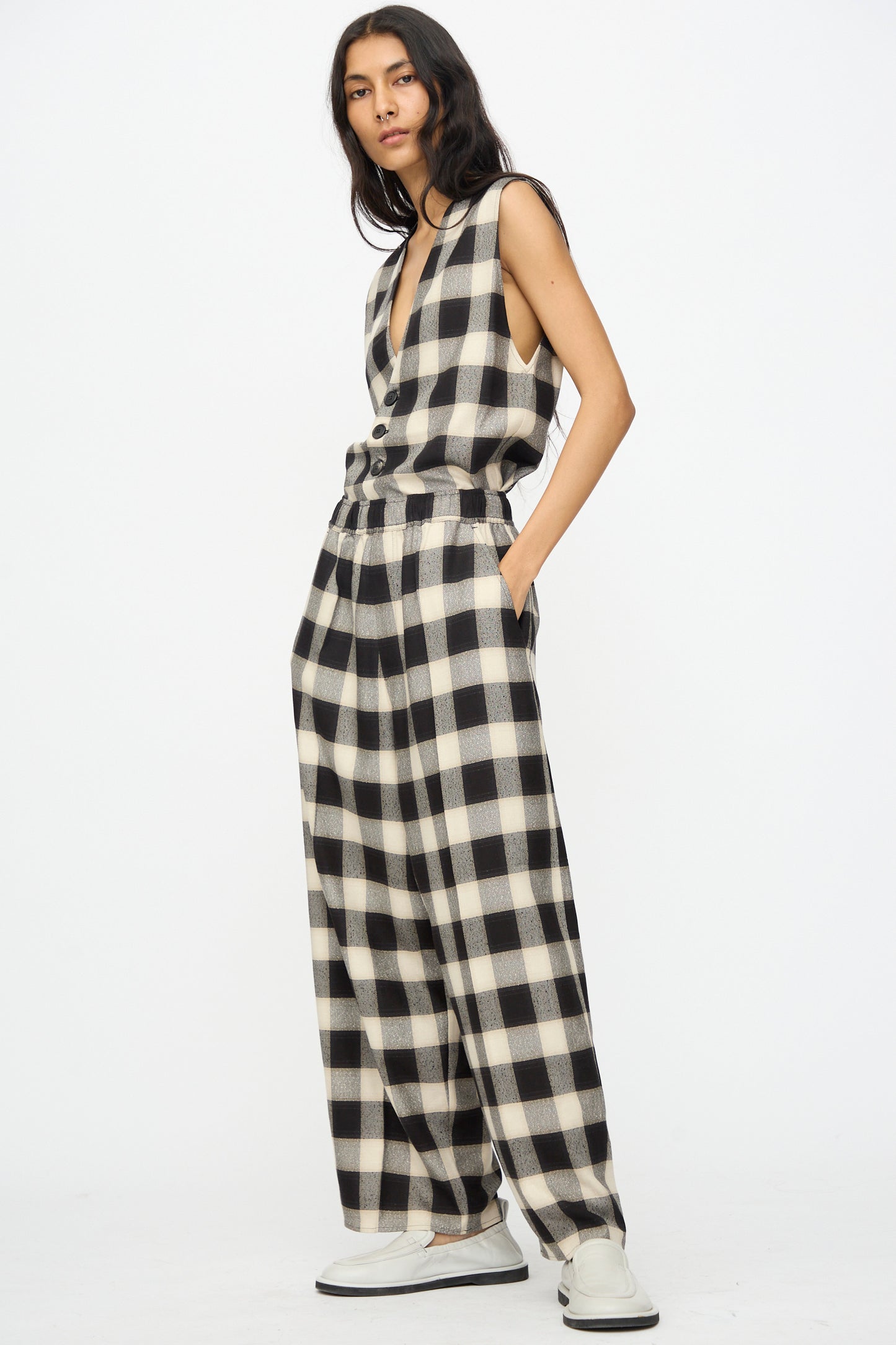 A person stands against a white background wearing Cristaseya's Oversized Pyjama Pants in Black and Cream Checks with wide legs, paired with a sleeveless top and white shoes, hands casually in pockets.