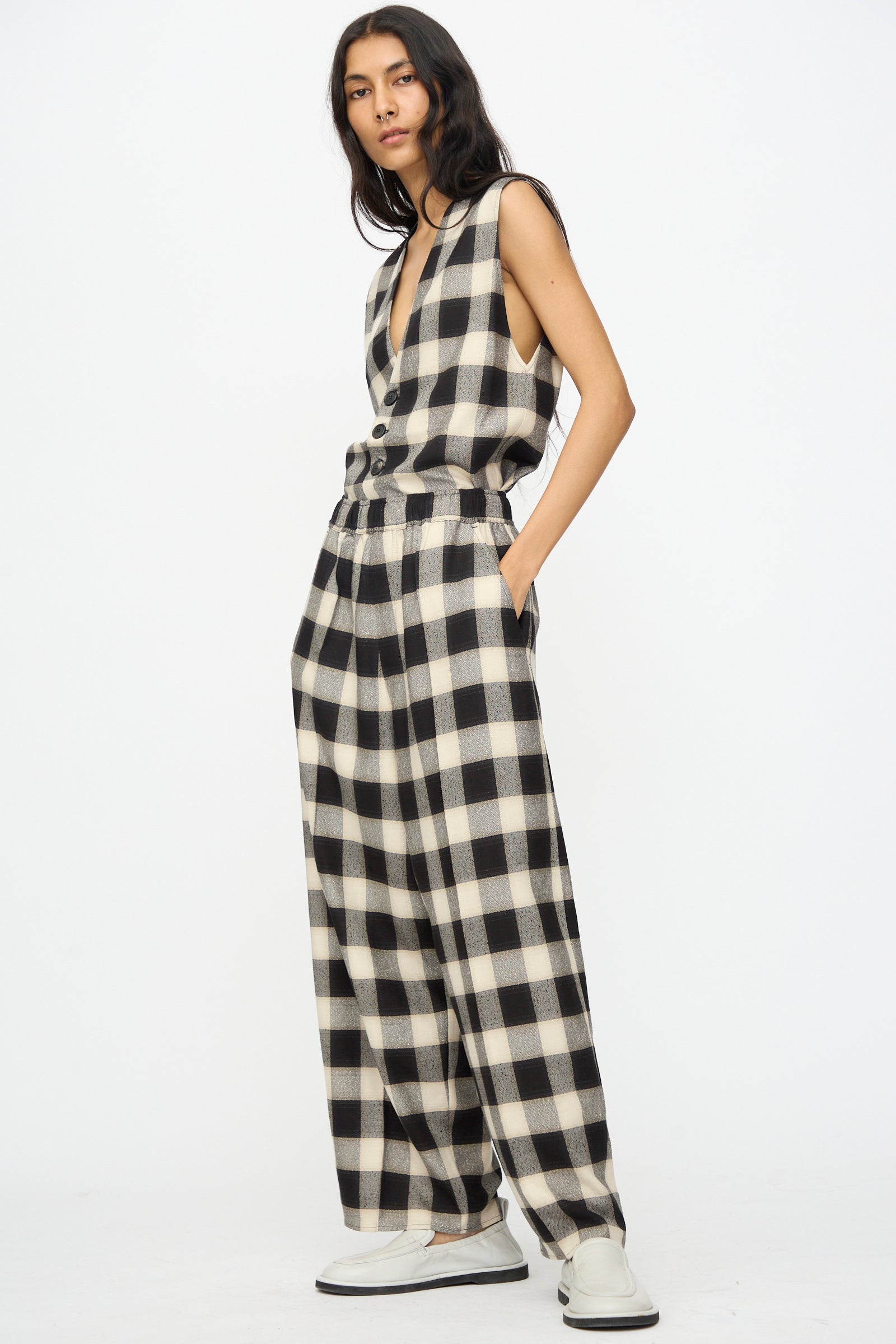 A person stands against a white background wearing Cristaseya's Oversized Pyjama Pants in Black and Cream Checks with wide legs, paired with a sleeveless top and white shoes, hands casually in pockets.