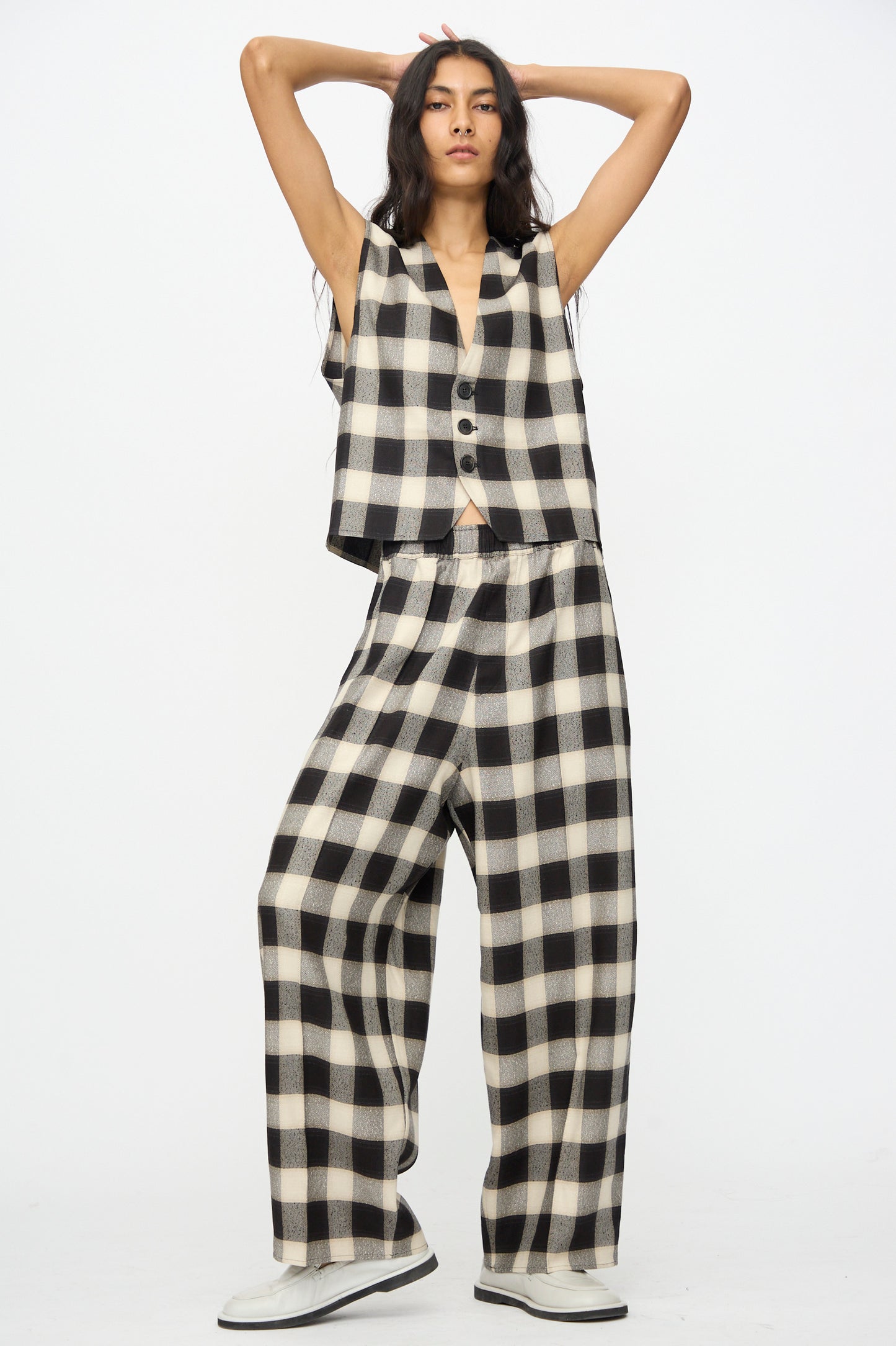 A woman stands against a white background wearing Cristaseya’s Oversized Pyjama Pants in Black and Cream Checks with a matching vest and white shoes.