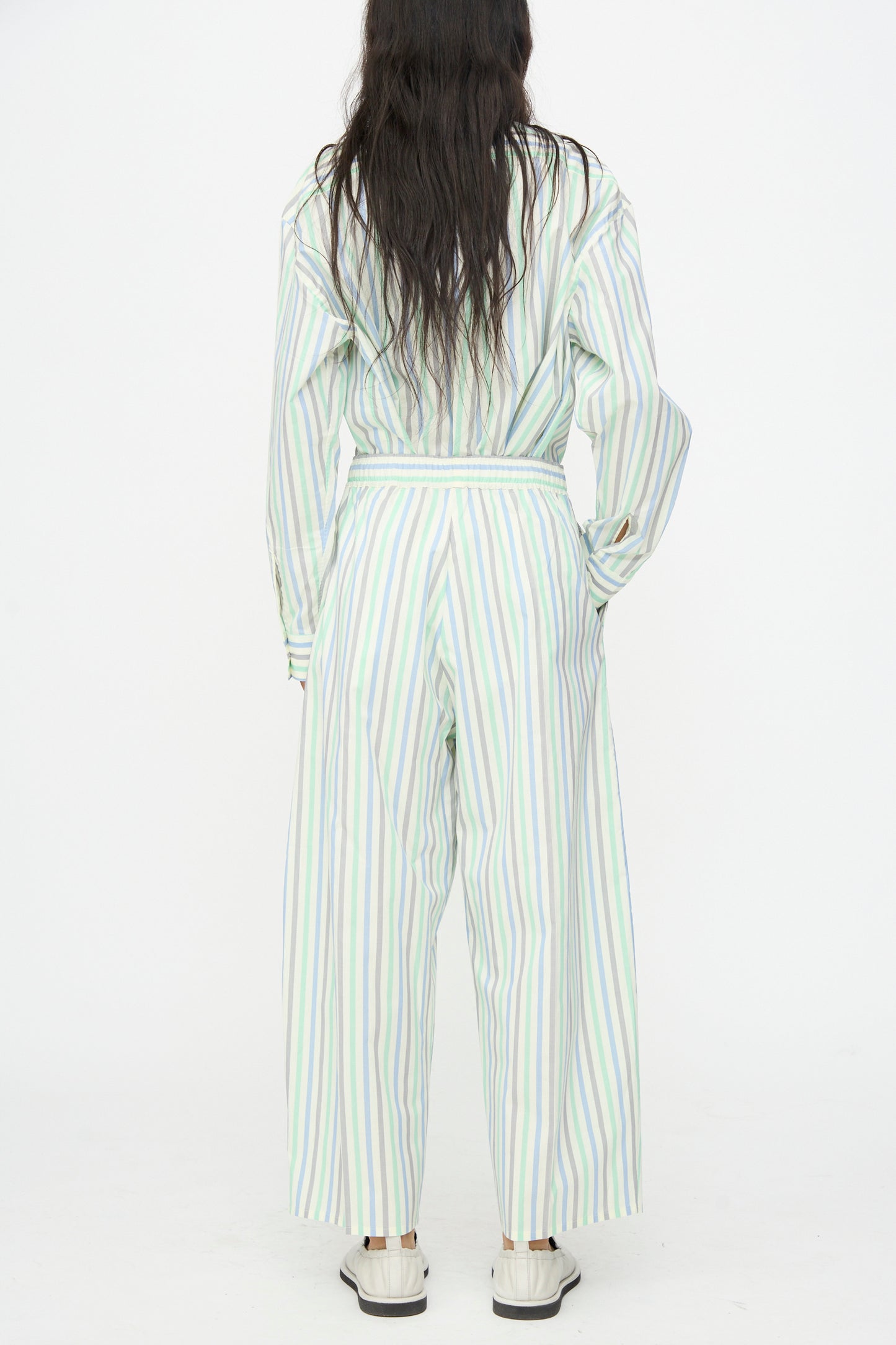 Person stands with back to camera, wearing Cristaseya Oversized Pyjama Pants in Striped Pastel Green and white sneakers, against a plain white background.
