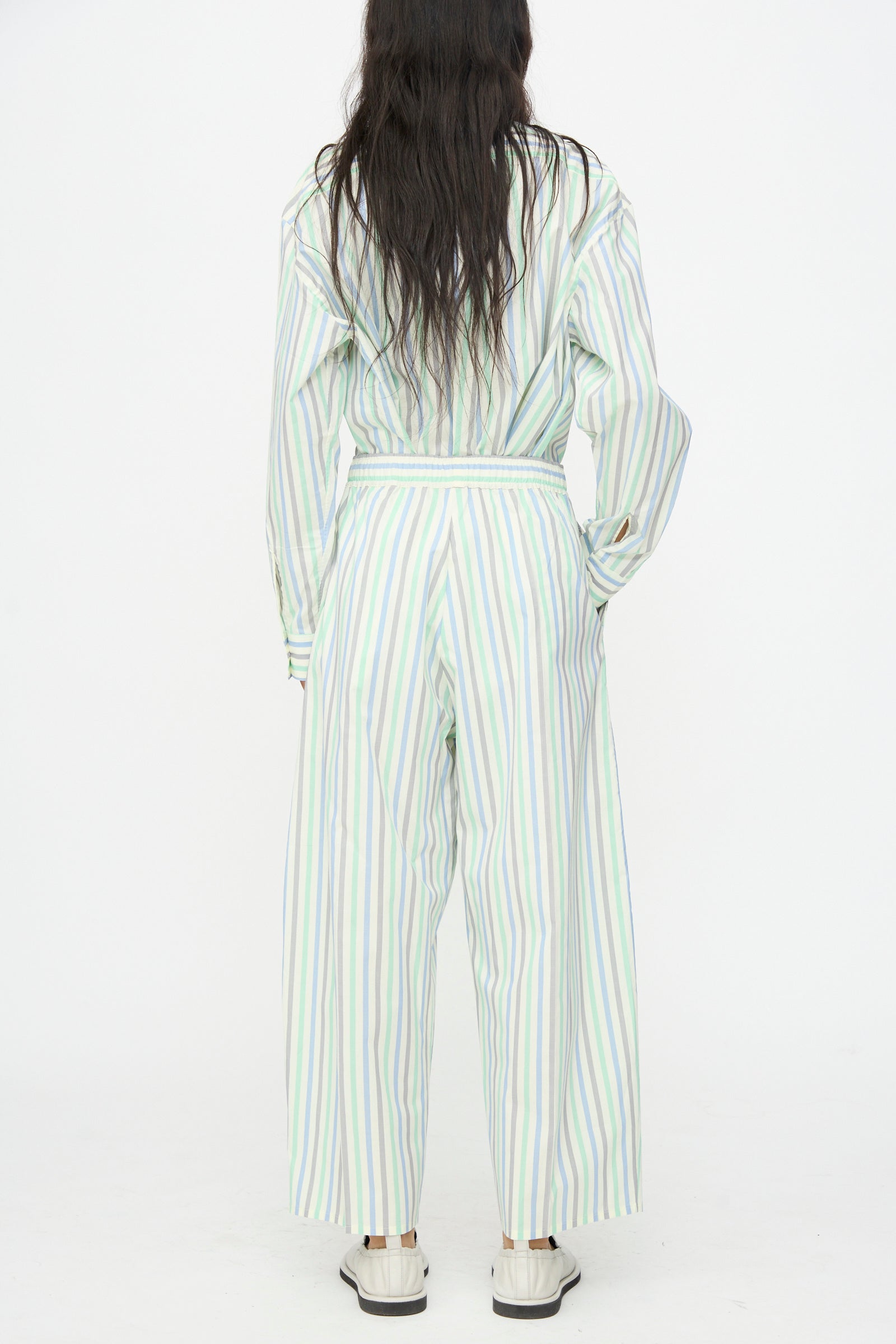 Person stands with back to camera, wearing Cristaseya Oversized Pyjama Pants in Striped Pastel Green and white sneakers, against a plain white background.
