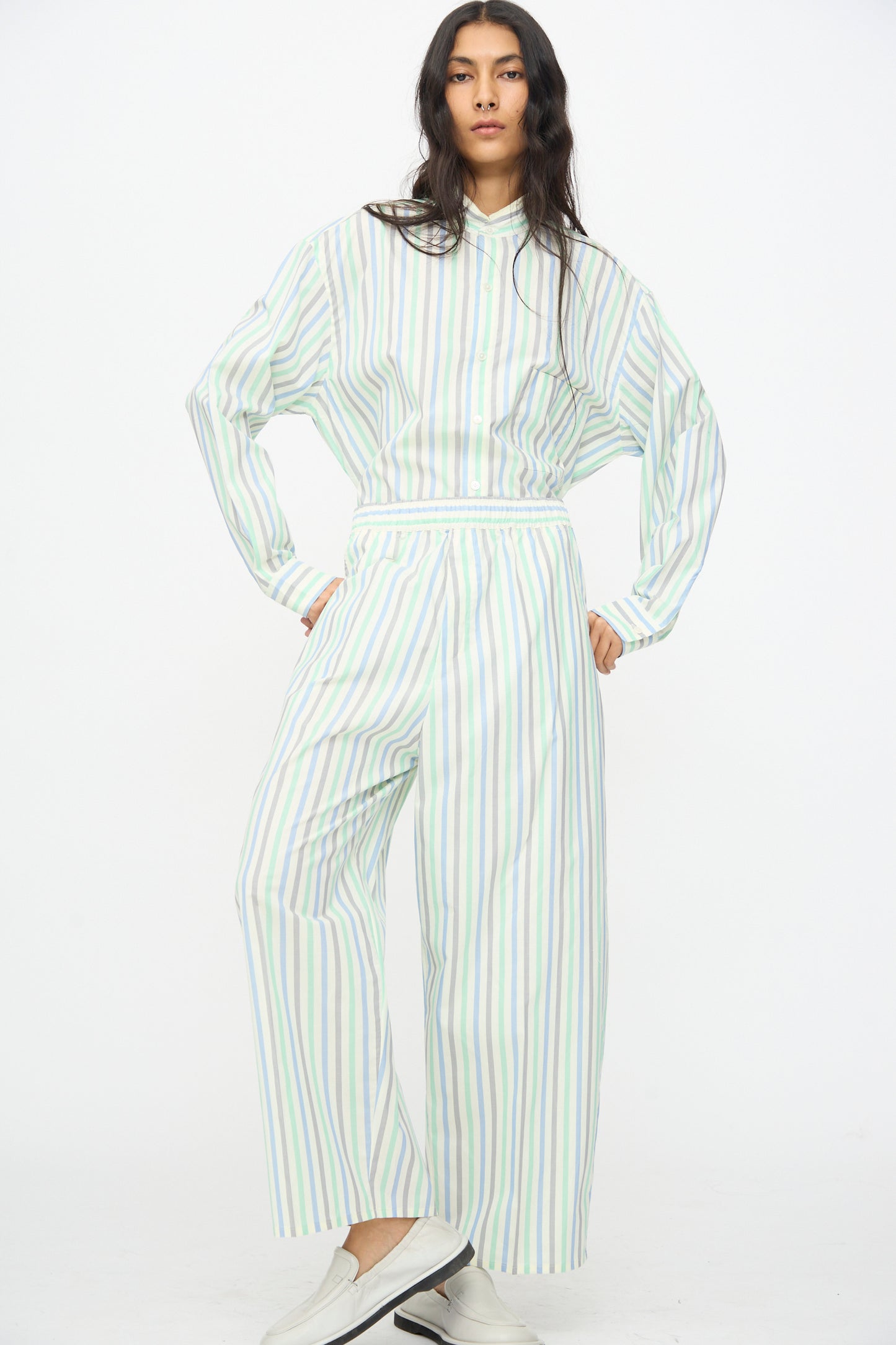 A person stands against a white background, wearing Cristaseya Oversized Pyjama Pants in Striped Pastel Green styled with a matching loose shirt and white shoes.