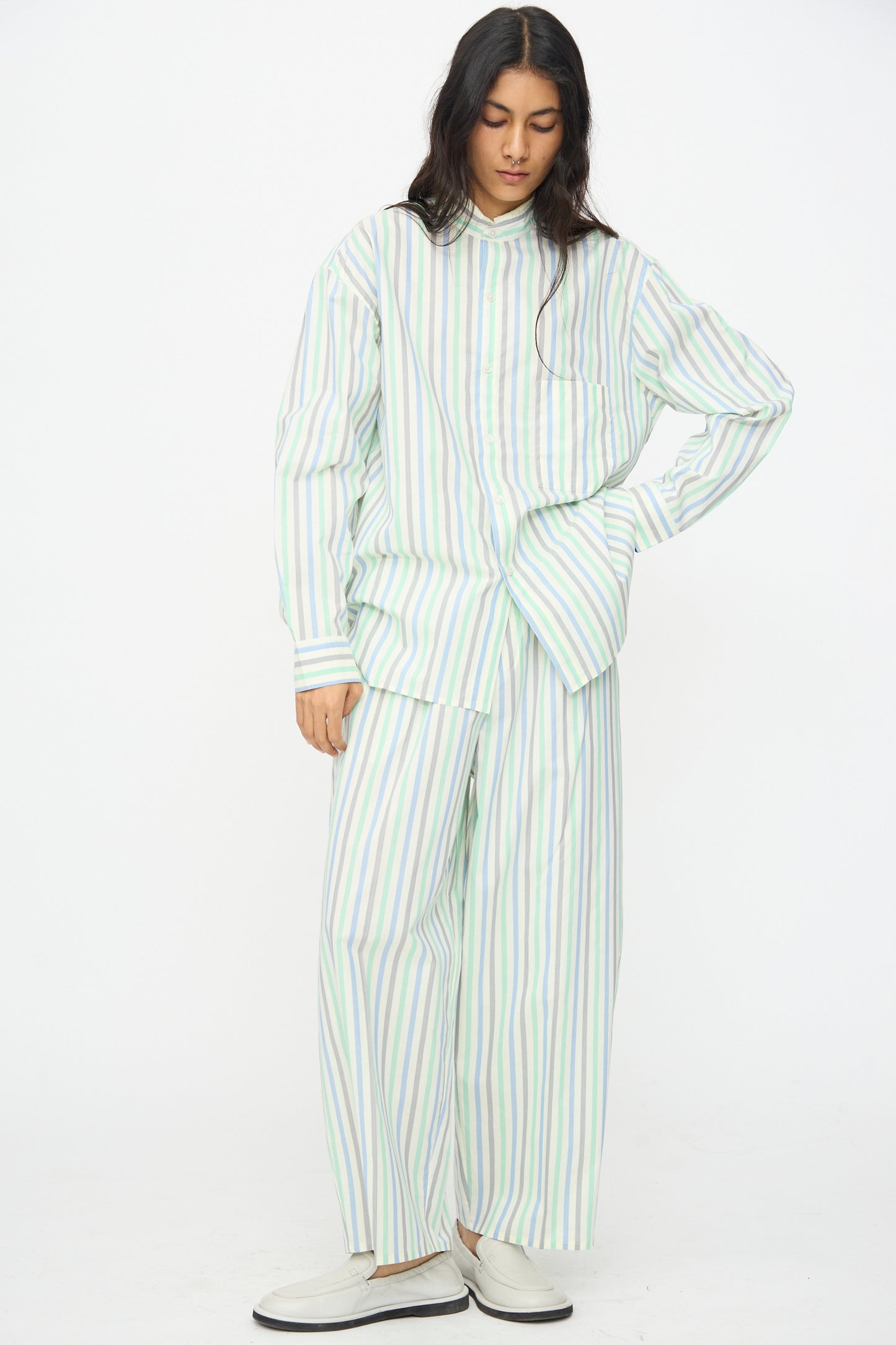 A person stands against a white background in a loose shirt, Cristaseya Oversized Pyjama Pants in Striped Pastel Green, and white shoes.