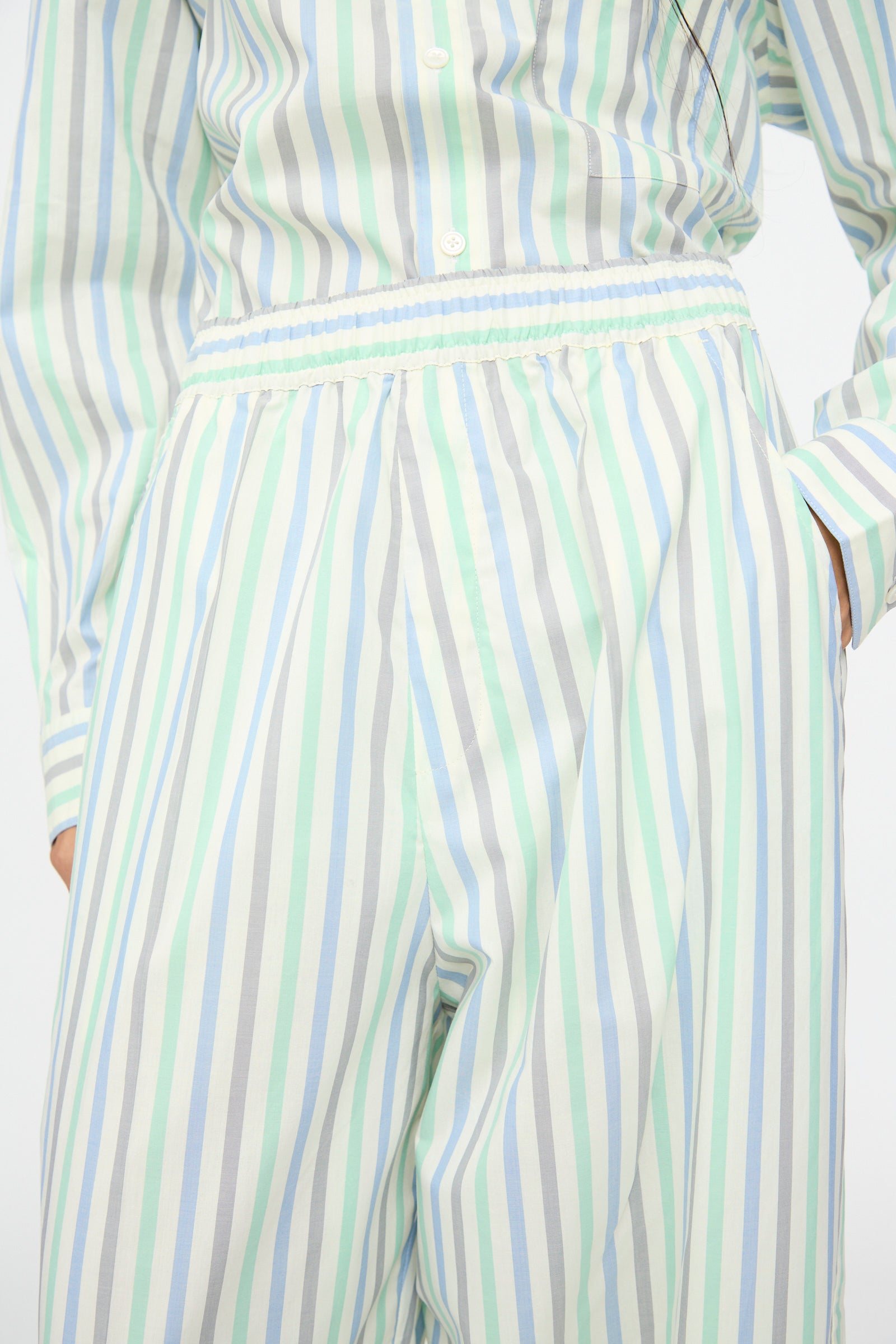A person wearing Cristaseya's Oversized Pyjama Pants in Striped Pastel Green, paired with a matching oversized top, both featuring light blue, green, and white vertical stripes, with hands partly in pockets.