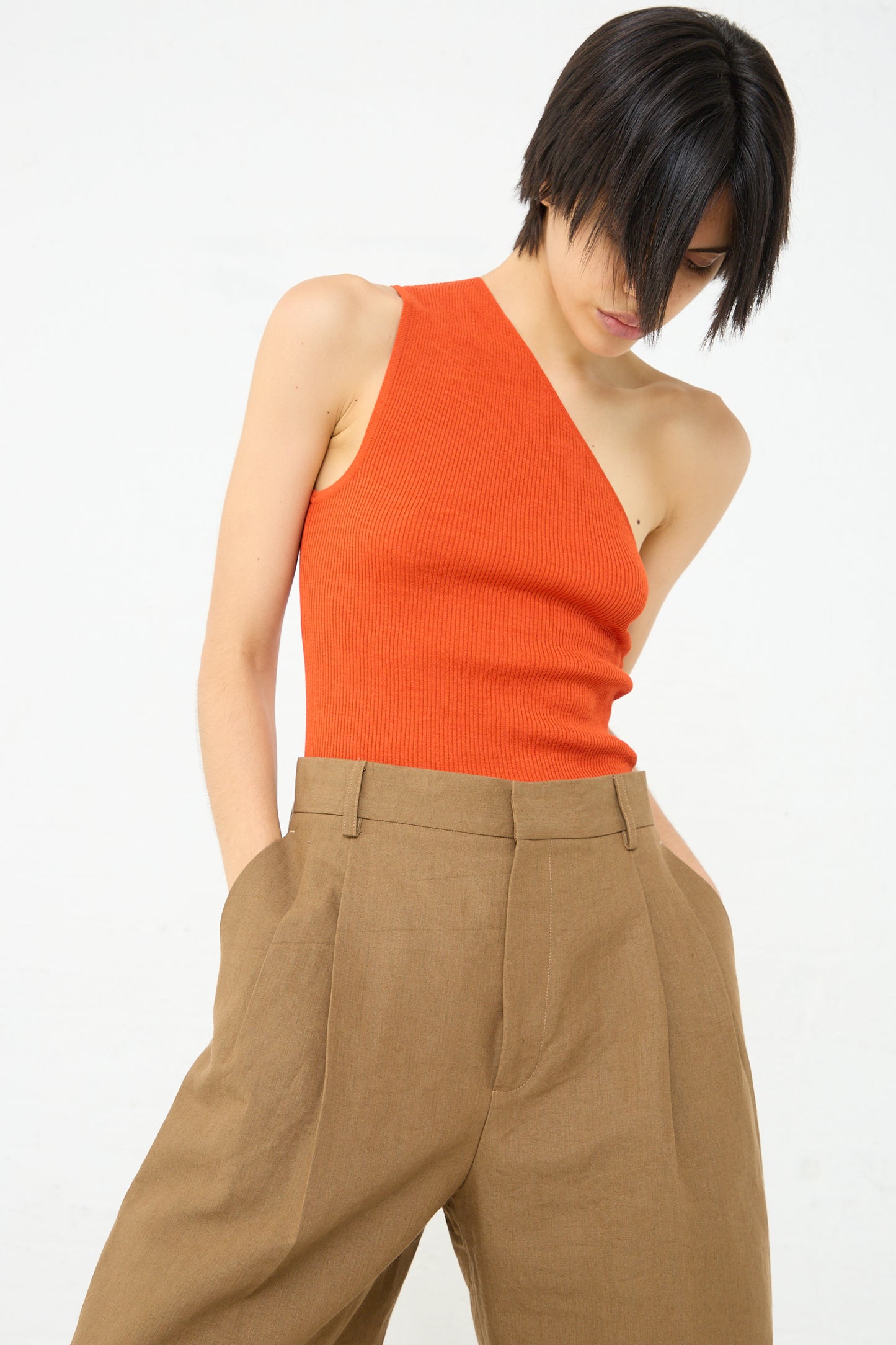 A woman in a Cristaseya Ribbed Silk Asymmetrical Top in Orange with a one-shoulder design and tan pants.