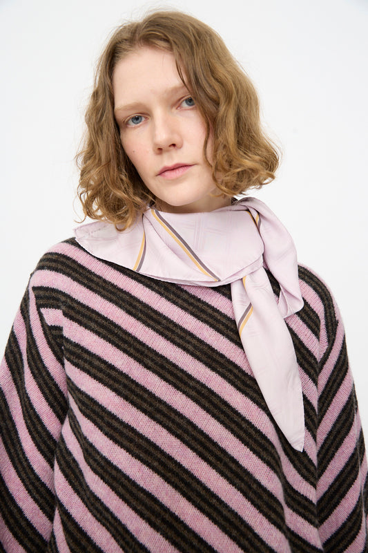 A woman with wavy, shoulder-length hair stands against a plain white background, wearing a black and pink striped sweater and the Cristaseya Silk Carré in Light Pink Checks tied around her neck.