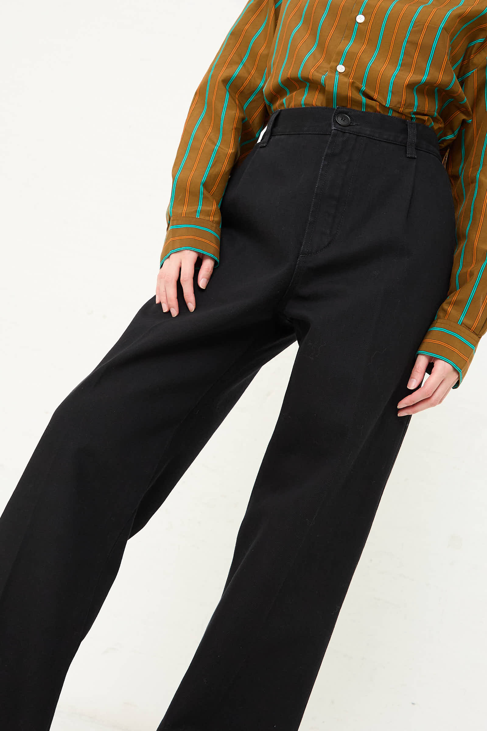 A person wearing a brown and green striped shirt is seen in the image, paired with Cristaseya's One Wash Denim Pleated Trouser in Black. The high-waisted pants complement the casual style, capturing the torso and legs as the individual slightly leans left.