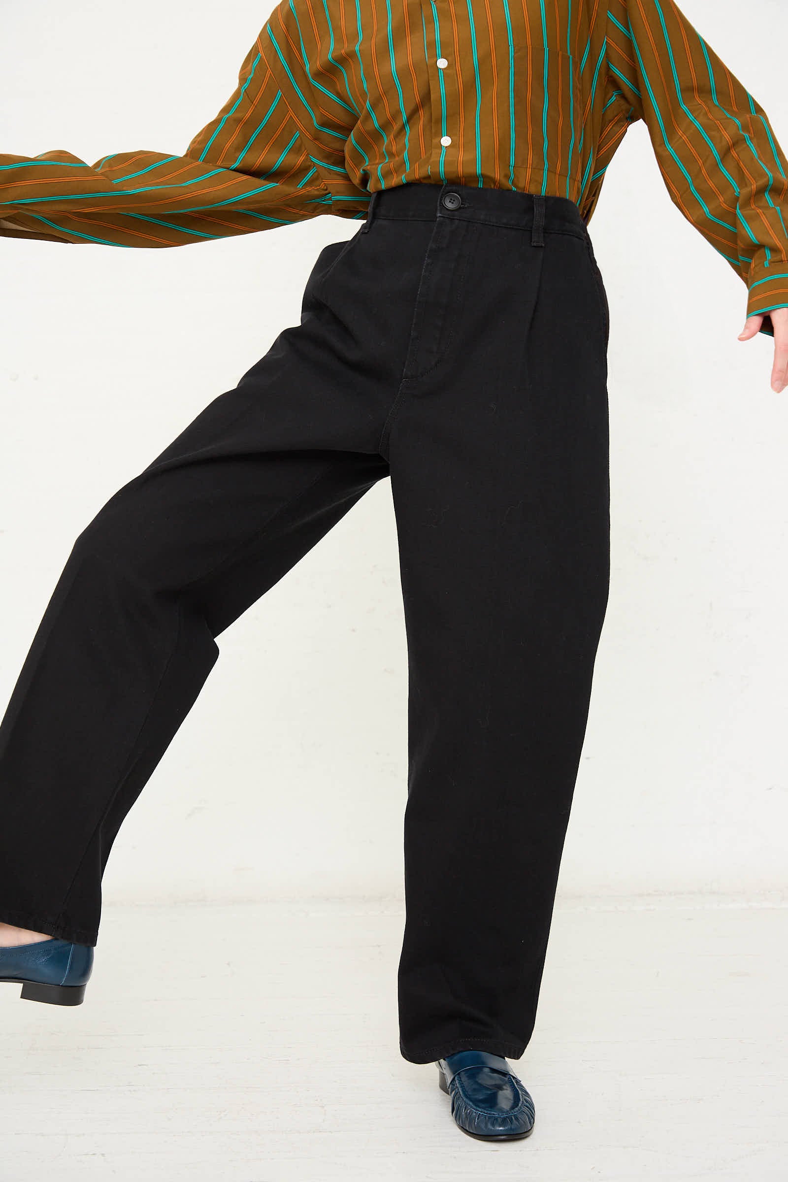 A person wearing a striped brown and green shirt with Cristaseya One Wash Denim Pleated Trousers in Black and dark shoes, poses against a plain light background.