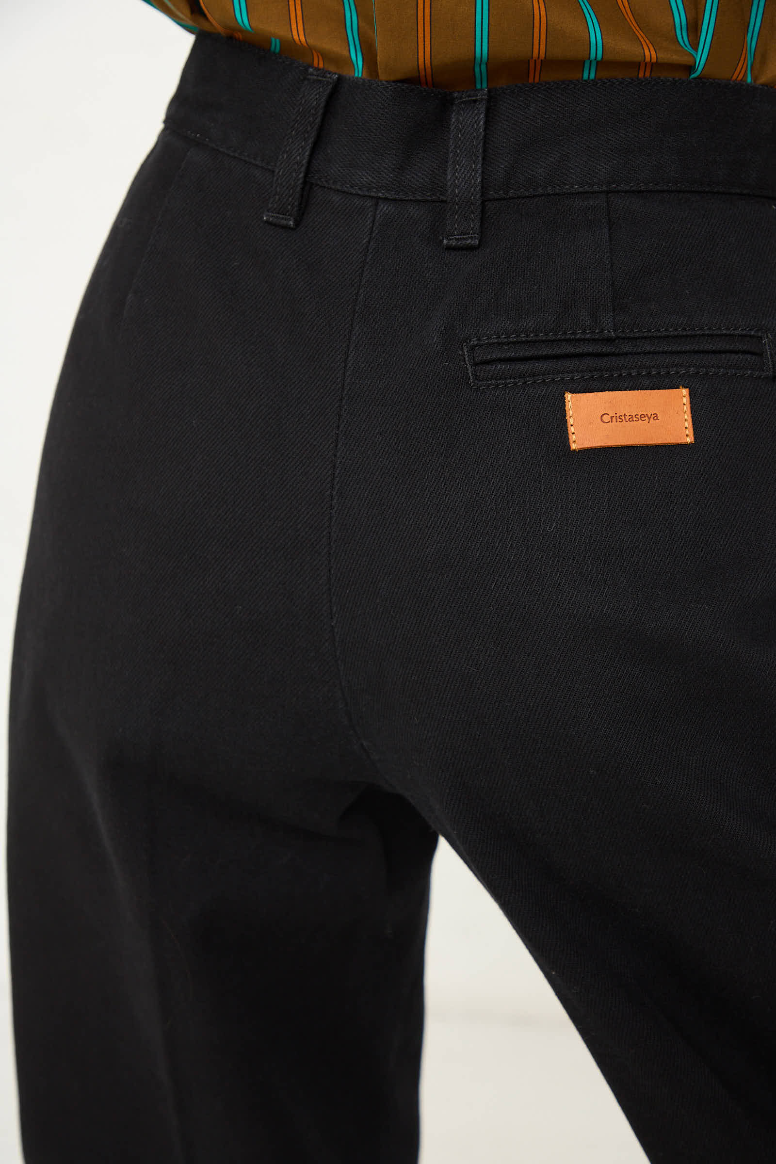 Close-up of a person wearing the One Wash Denim Pleated Trouser in Black with a brown striped shirt tucked in. A small brown label reading "Cristaseya" is seen on the back pocket, highlighting the relaxed fit and 100% cotton material.