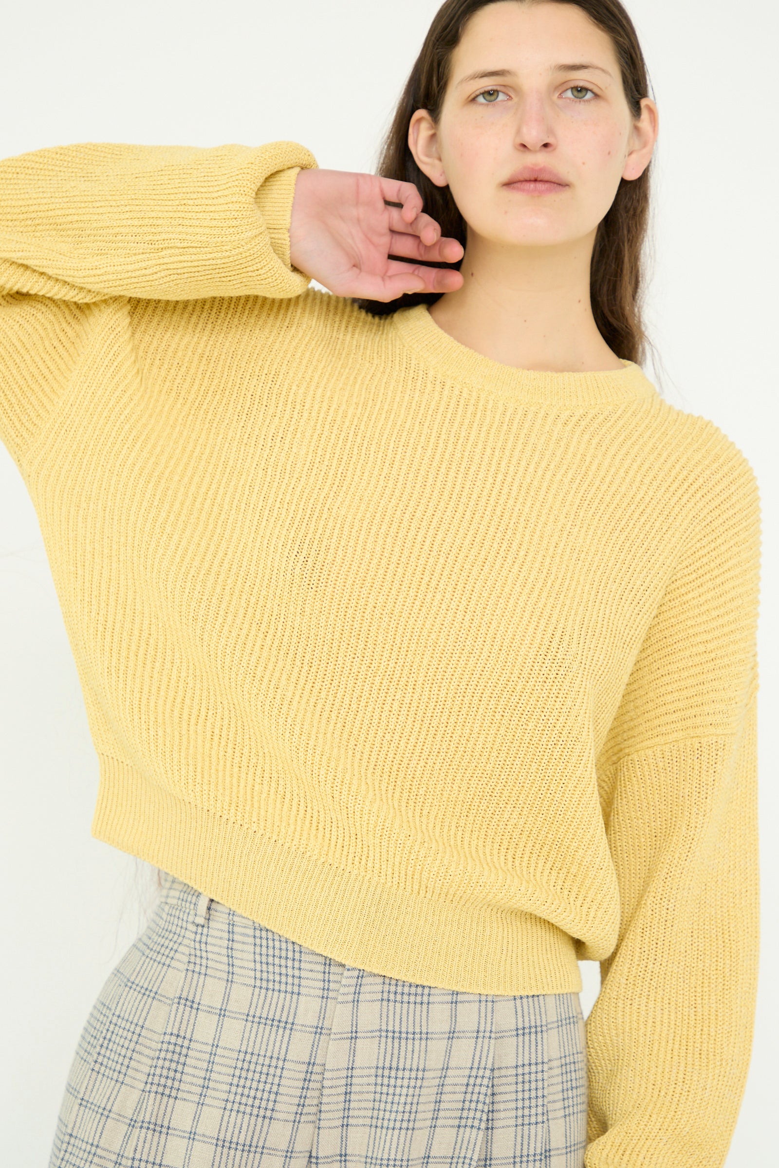 A person poses against a plain background, one hand next to their head, wearing plaid pants and a yellow Washi Paper Dropshoulder Cropped Sweater by Cristaseya.