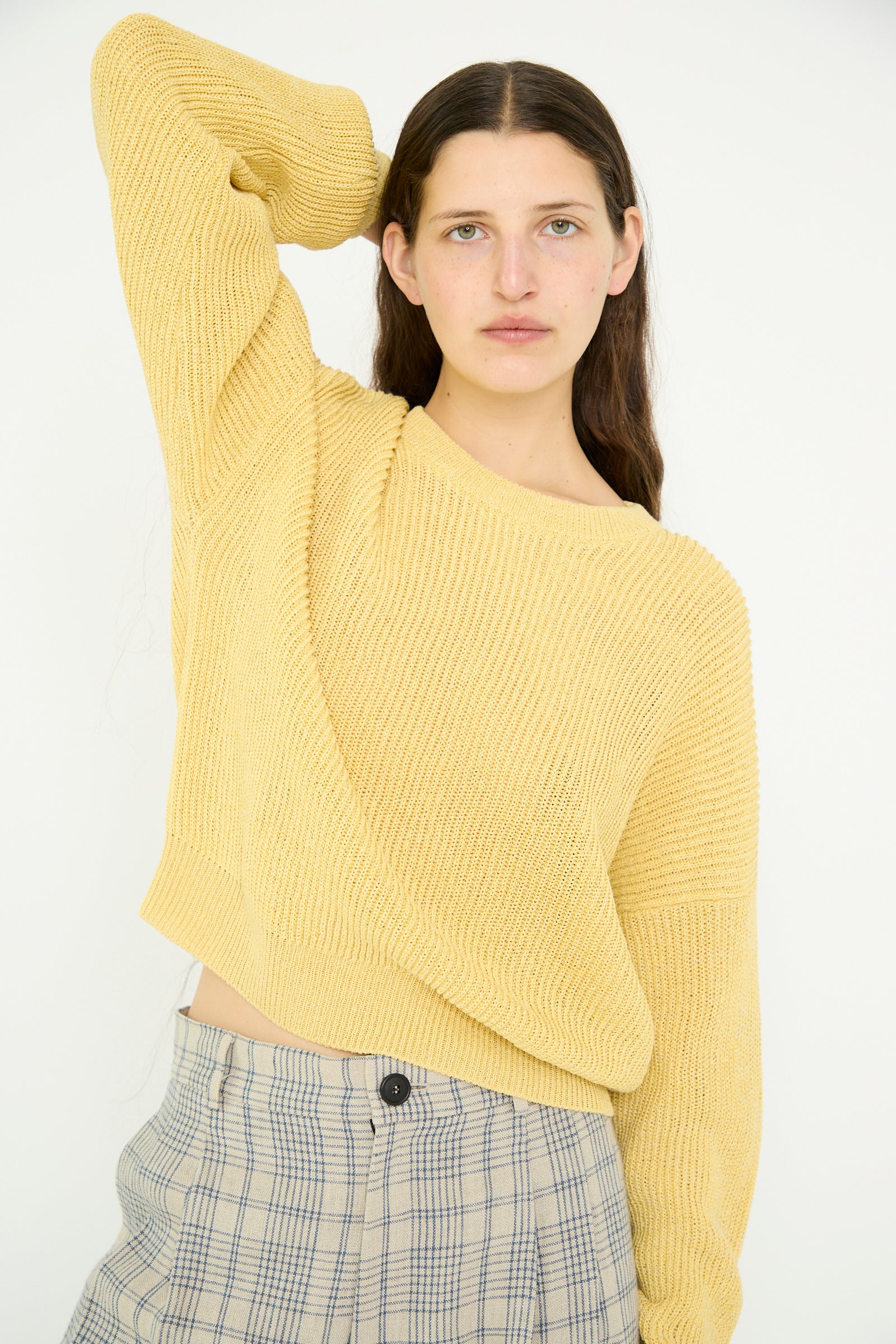 A model with long hair poses against a white background, one arm behind their head, wearing a Cristaseya Washi Paper Dropshoulder Cropped Sweater in Yellow and relaxed fit plaid pants.