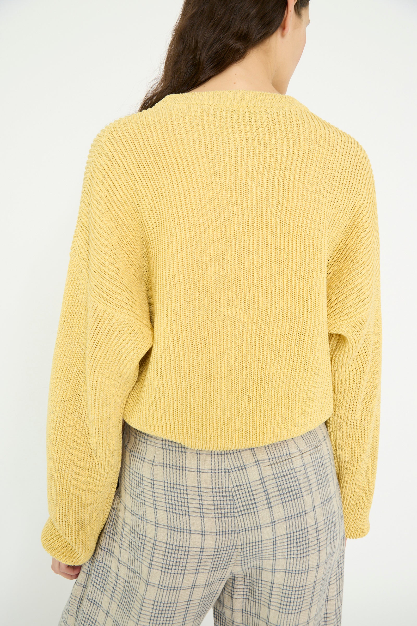 A person with long hair is seen from the back against a plain white background, wearing a Cristaseya Washi Paper Dropshoulder Cropped Sweater in Yellow and plaid pants.