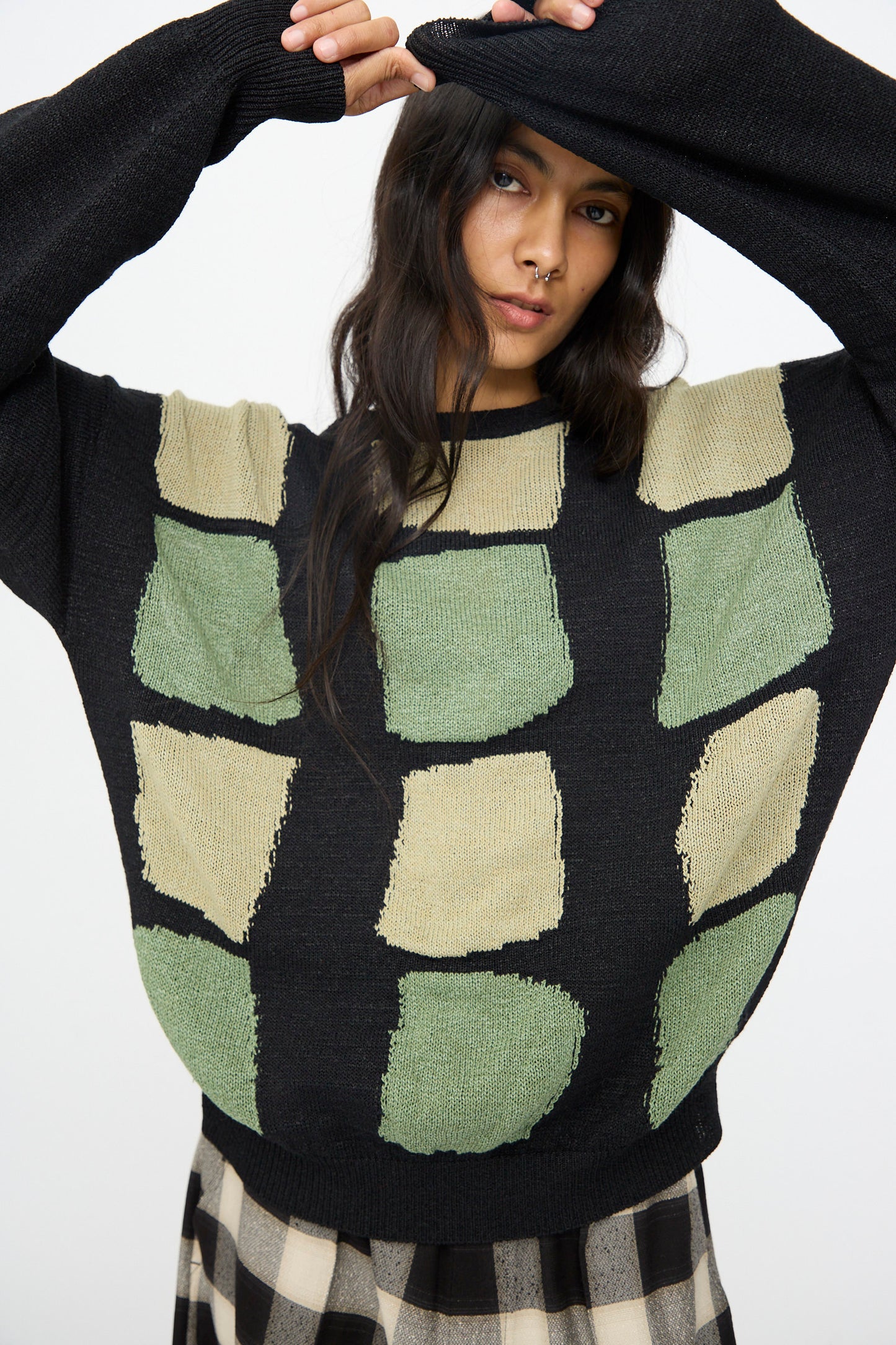 A person wearing the Cristaseya Washi Paper Patchwork Sweater in Black with Sage and Light Khaki stands with one arm raised, partially covering their head, paired with a black and beige checkered skirt.