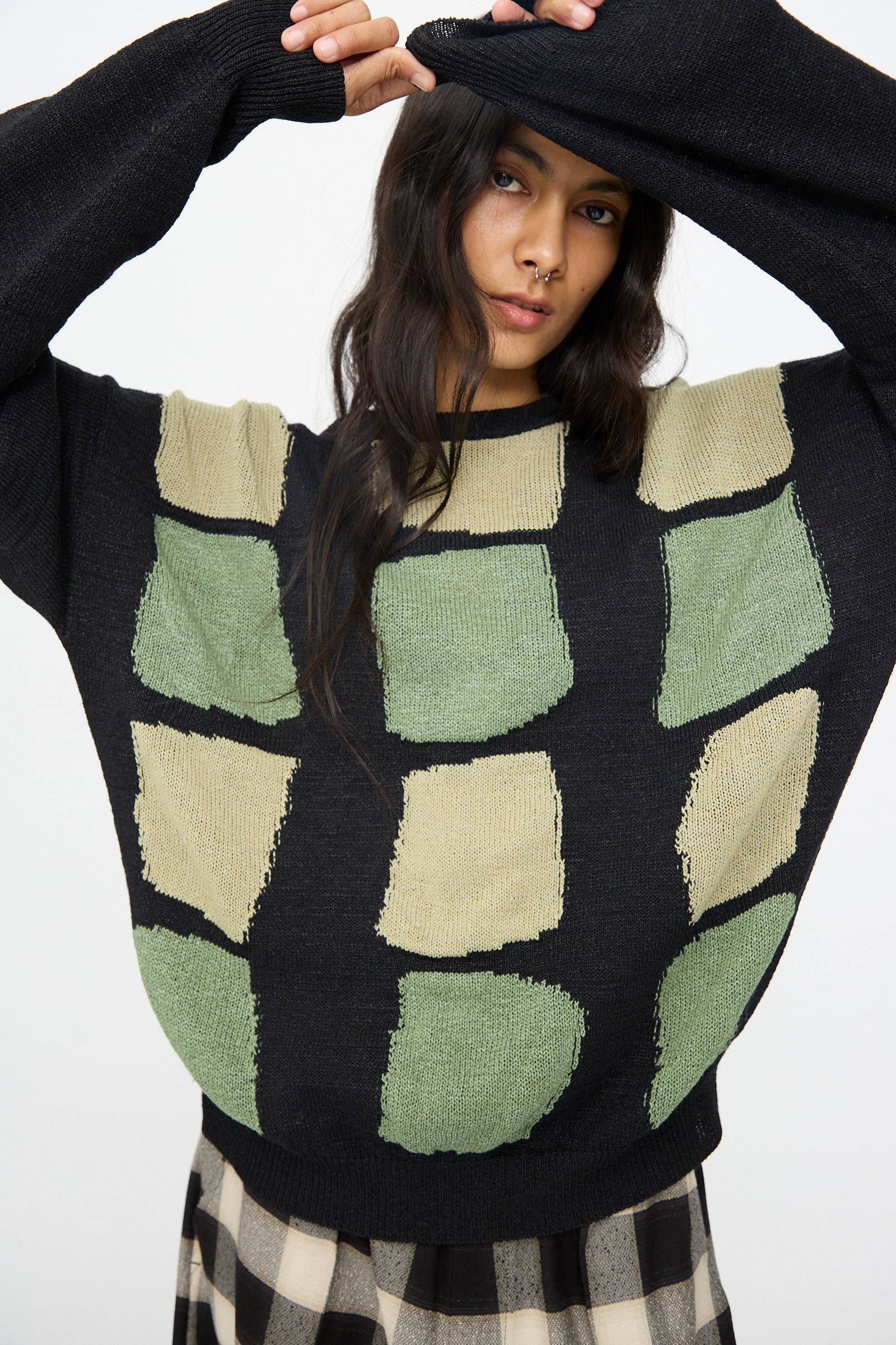 A person wearing the Cristaseya Washi Paper Patchwork Sweater in Black with Sage and Light Khaki stands with one arm raised, partially covering their head, paired with a black and beige checkered skirt.