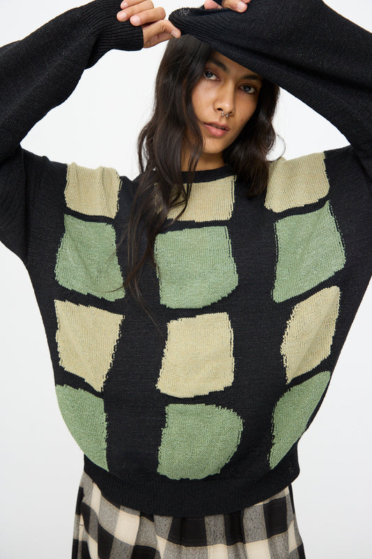A person wearing the Cristaseya Washi Paper Patchwork Sweater in Black with Sage and Light Khaki stands with one arm raised, partially covering their head, paired with a black and beige checkered skirt.
