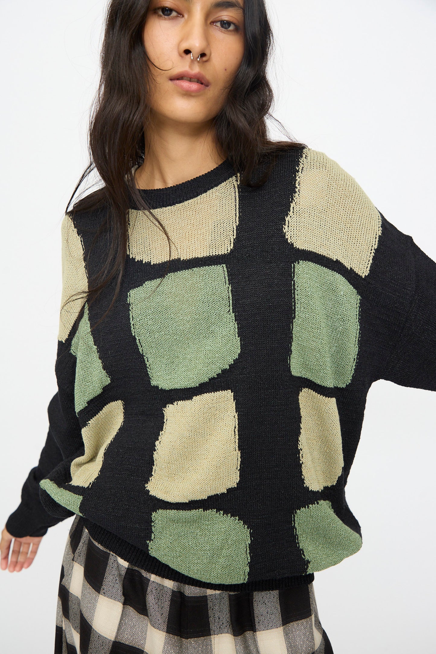A woman wears the Cristaseya Washi Paper Patchwork Sweater in Black with sage and light khaki squares, paired with a checkered skirt, standing against a plain white background.