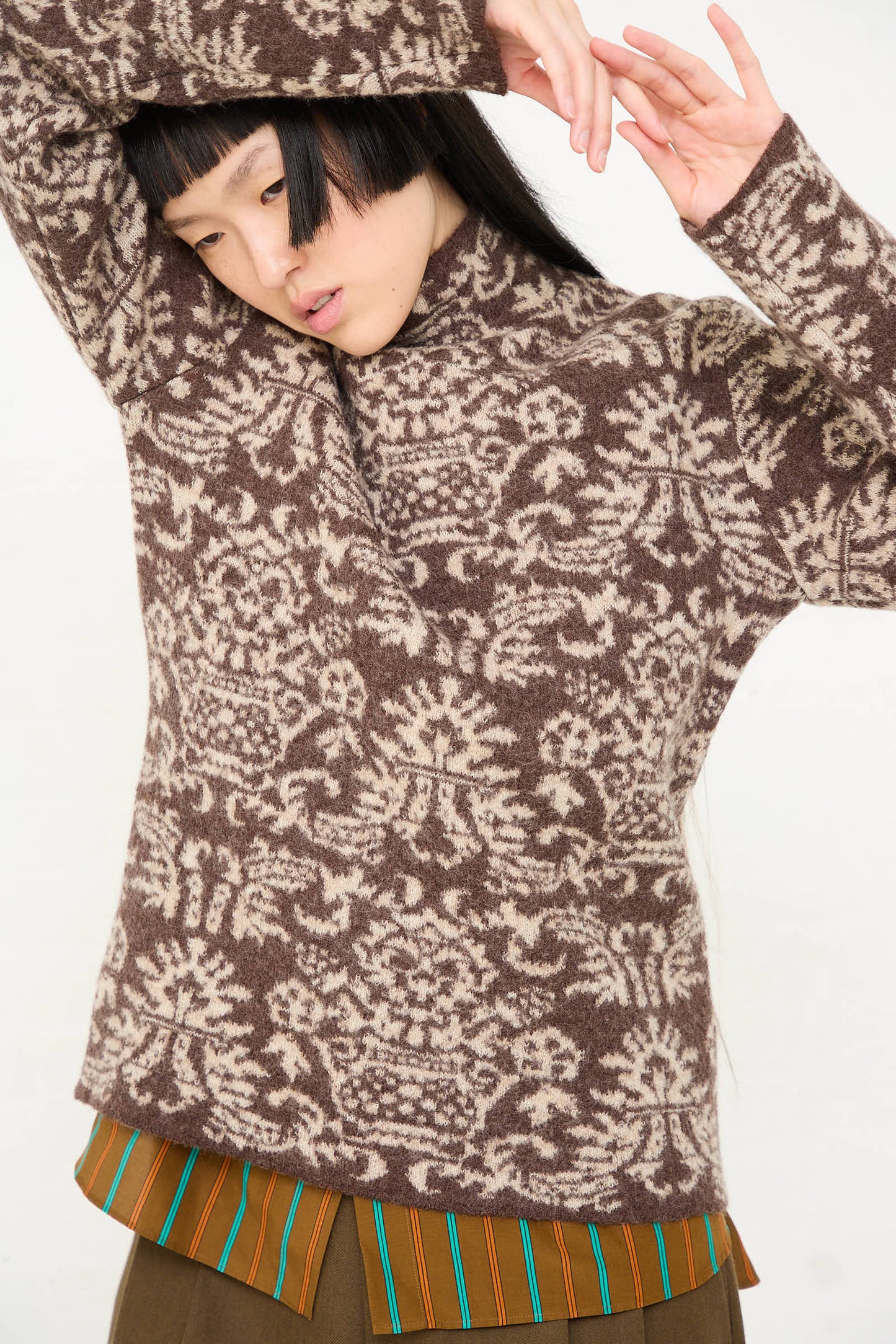 A person in a Cristaseya Wool Felted Jacquard Turtleneck Sweater in Mocha and Cream with arms raised is posing against a white background. They are also wearing a blouse with a striped collar and a brown skirt, showcasing an intricate jacquard pattern on the sweater.