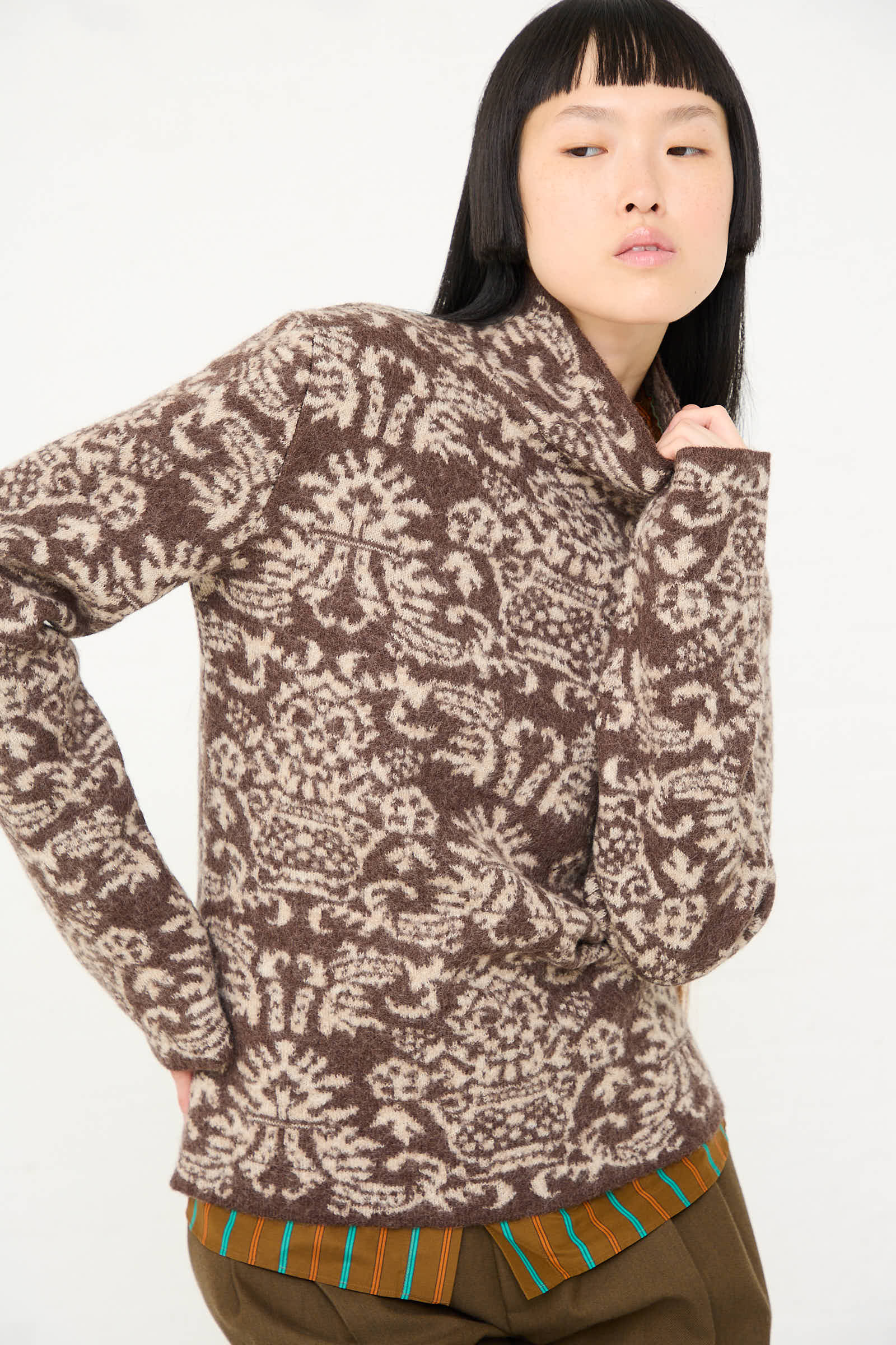 Person wearing a Wool Felted Jacquard Turtleneck Sweater in Mocha and Cream by Cristaseya over a striped shirt, posing with one hand behind their neck.