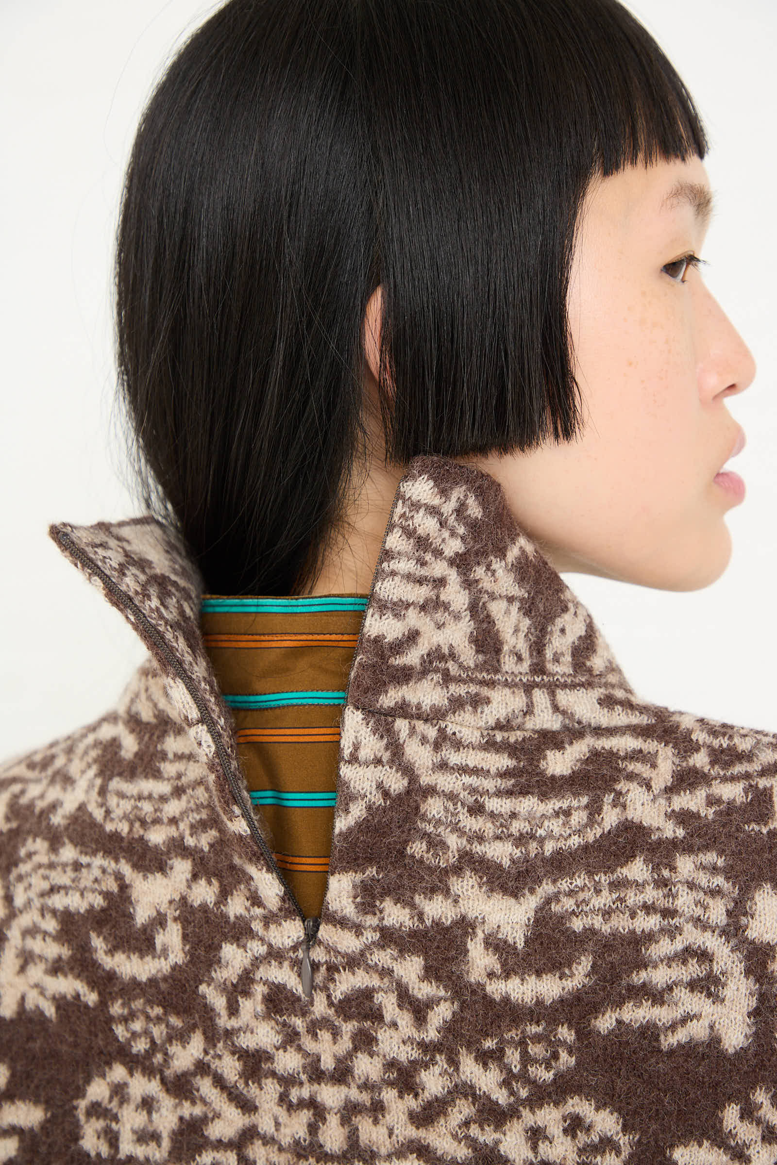 Profile view of a person with short black hair wearing the Cristaseya Wool Felted Jacquard Turtleneck Sweater in Mocha and Cream, layered over a striped brown and blue shirt.