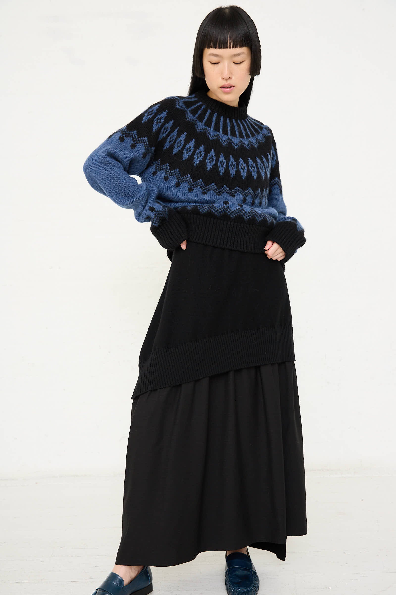 A person stands against a white background wearing a blue patterned wool sweater and the Wool Patched Long Skirt in Black by Cristaseya. They have a straight hairstyle with bangs and are looking slightly downward.