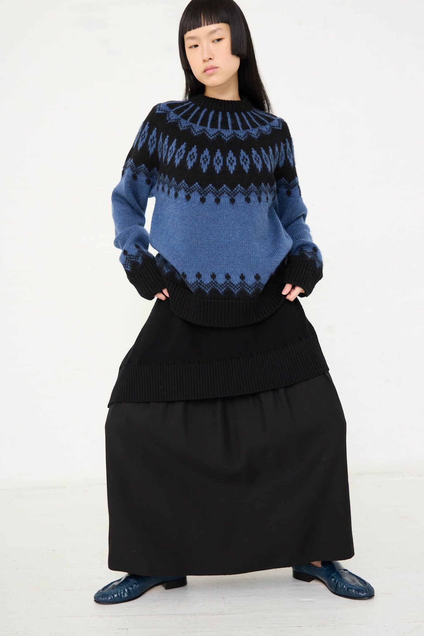 A person is standing against a plain background, wearing a blue and black patterned wool sweater, Cristaseya's Wool Patched Long Skirt in Black, and blue shoes.