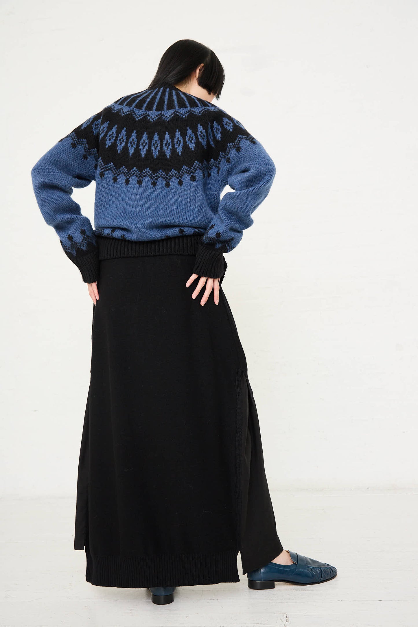 A person is standing with their back to the camera, wearing a blue and black patterned sweater, the Cristaseya Wool Patched Long Skirt in Black made in Italy, and blue shoes.