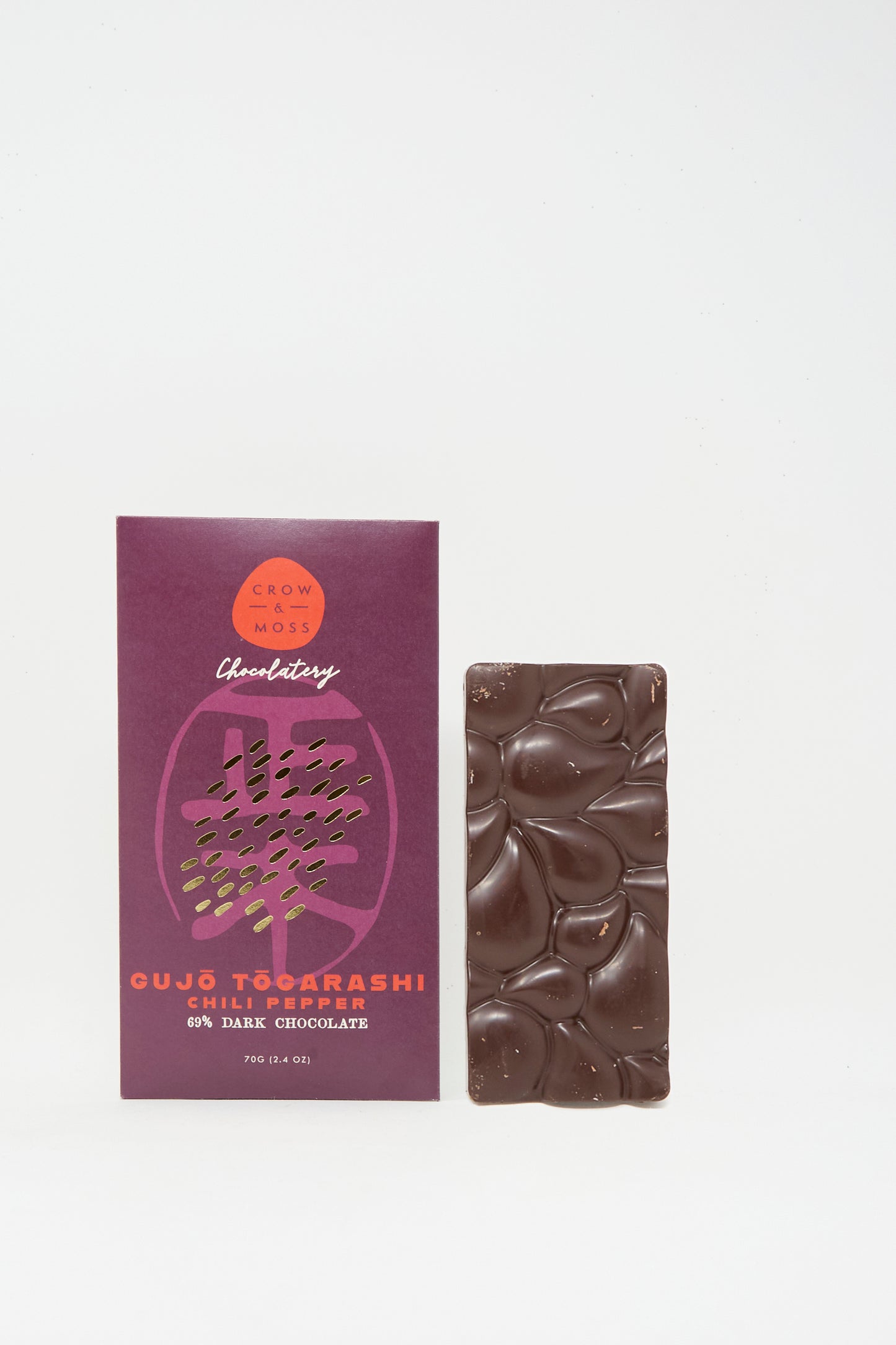 A textured bar of dark chocolate sits beside a purple Crow & Moss box labeled "Togarashi Chili Dark Chocolate Bar," crafted with single-origin Wampusirpi cacao for a unique, limited-edition treat.