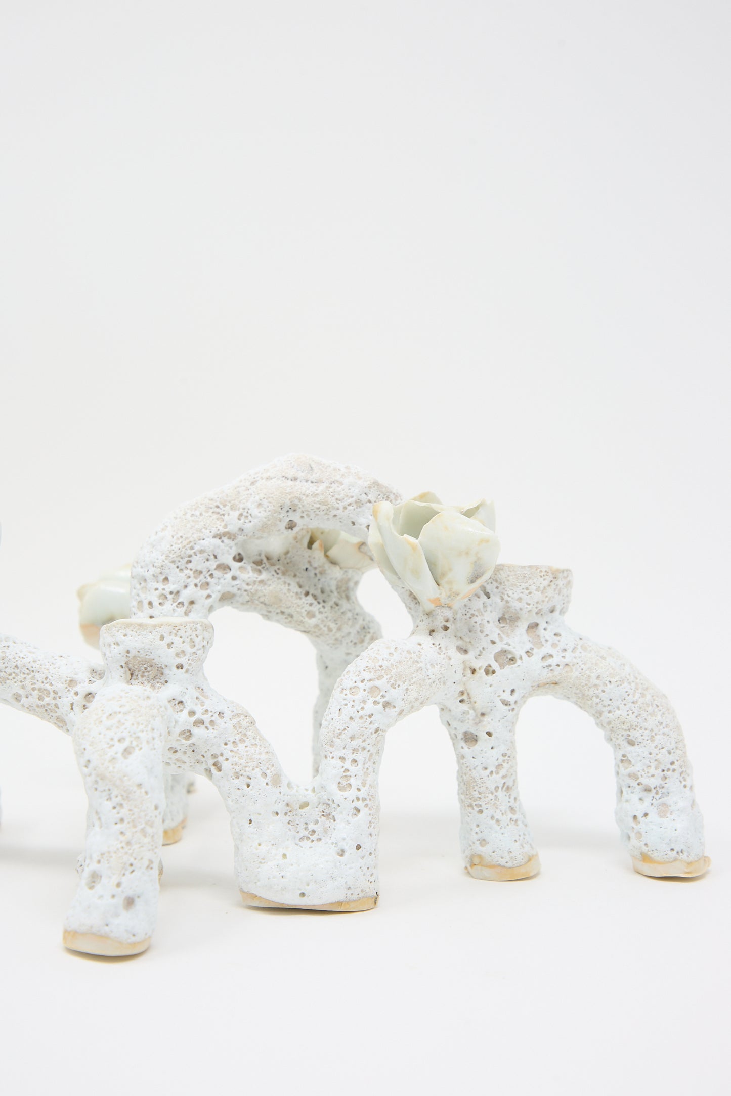 White, porous ceramic sculpture with an abstract, organic design featuring arch-like shapes and small, protruding elements. One of the arches has a flower-like detail on top, resembling luminescent petals. This hand-built piece stands against a plain white background. The **Blooming Arches Candle Holder** by **Dear You** beautifully accentuates any space with its intricate craftsmanship.