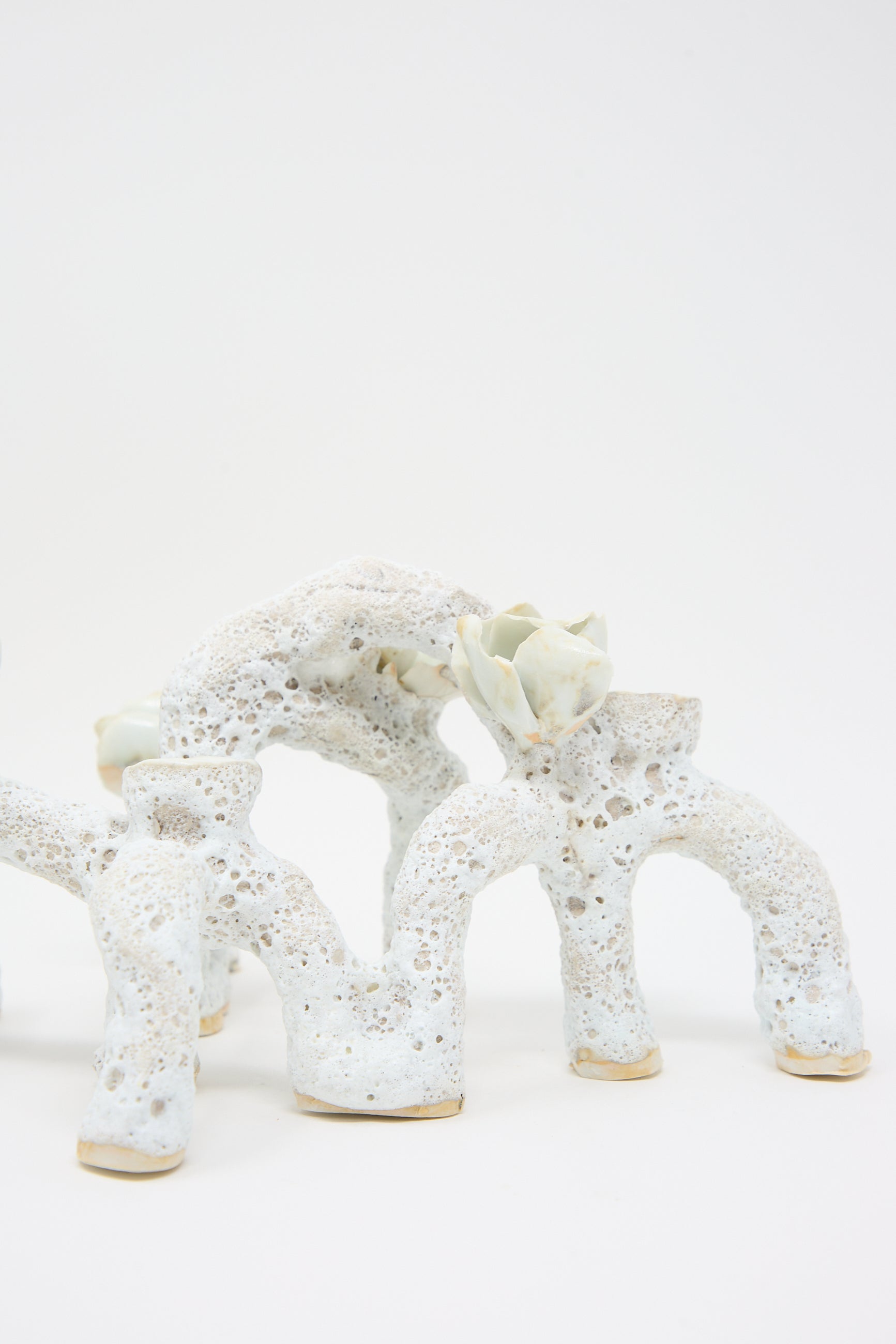 White, porous ceramic sculpture with an abstract, organic design featuring arch-like shapes and small, protruding elements. One of the arches has a flower-like detail on top, resembling luminescent petals. This hand-built piece stands against a plain white background. The **Blooming Arches Candle Holder** by **Dear You** beautifully accentuates any space with its intricate craftsmanship.