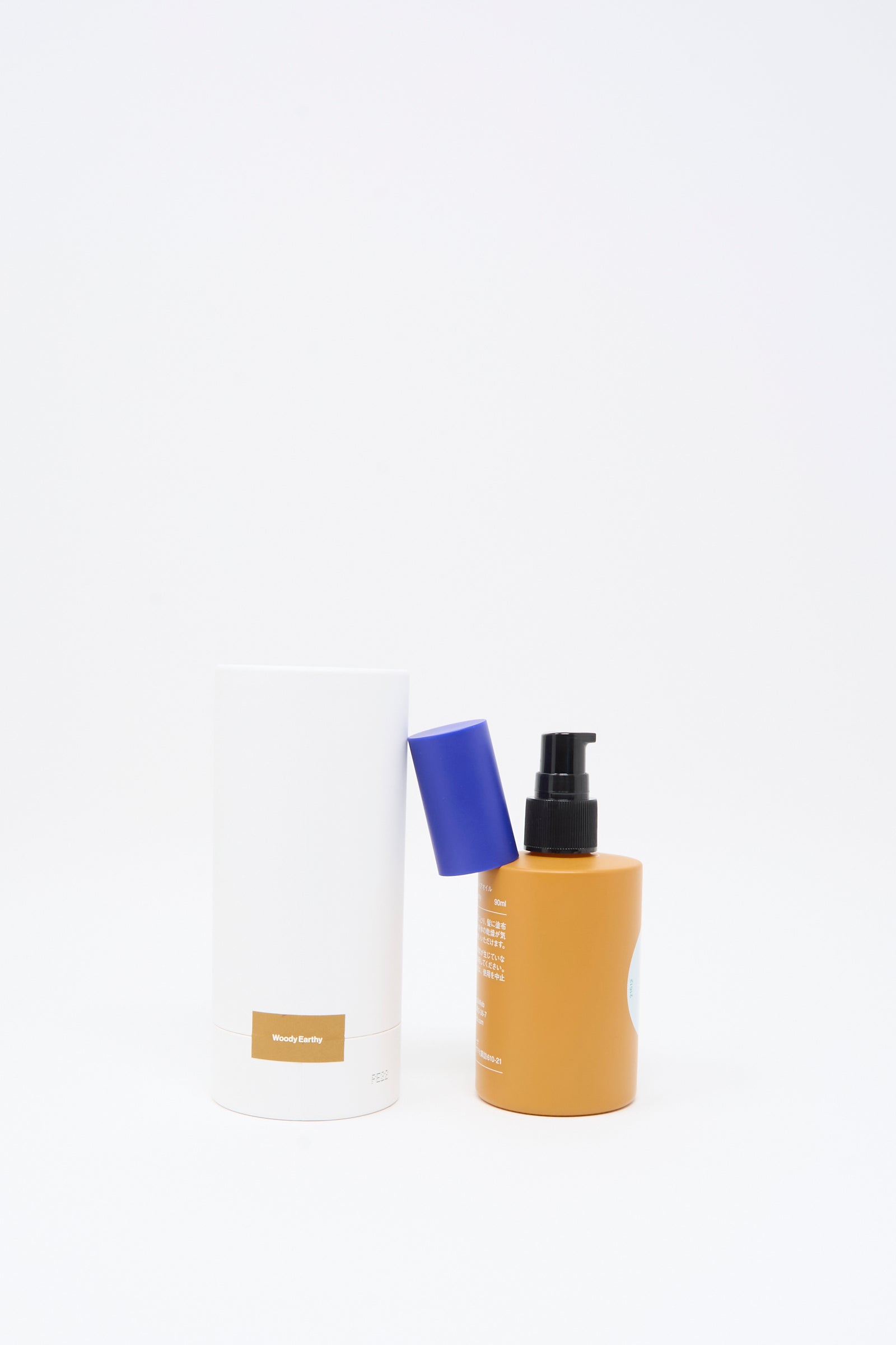 A white cylindrical container with a gold label stands next to Diffar's Woody Earthy Fragrance Oil, which comes in an amber pump bottle with its blue cap resting against the white container, all set against a plain white background.