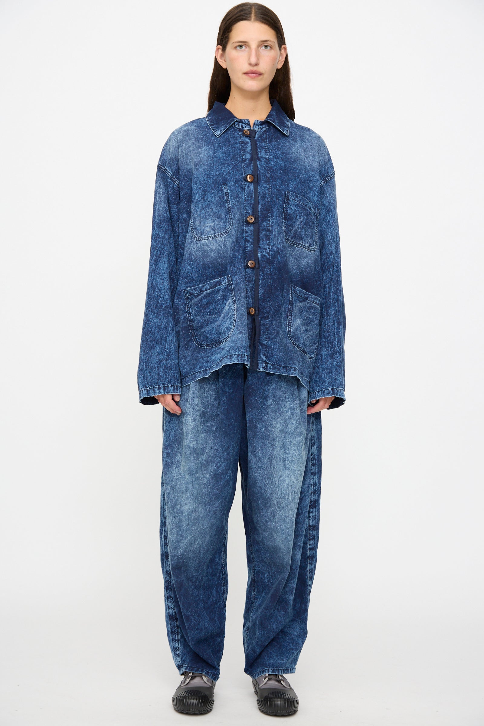 A person stands against a plain background wearing the Dr. Collectors 6 oz. Cotton New Saigon Shirt Jacket in Indigo—an oversized workwear style made in California—paired with loose-fitting matching jeans and black shoes.