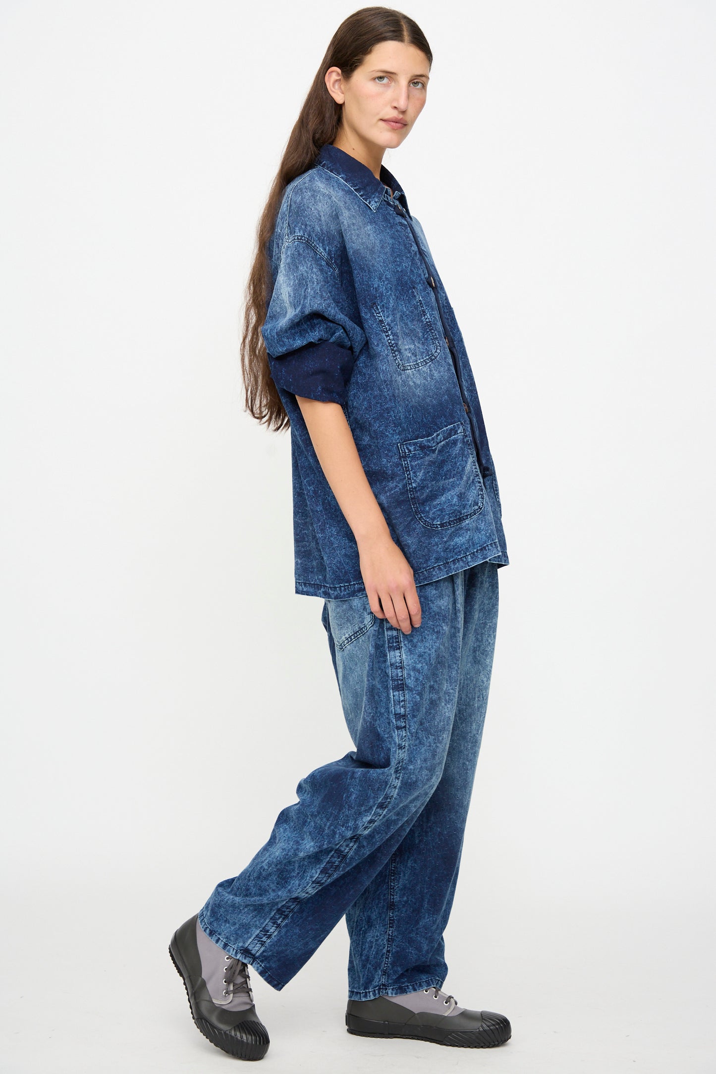 A person with long brown hair wears a loose-fitting, dark blue denim jacket and Dr. Collectors 6 oz. Cotton P43 Kyoto Pant in Sunfaded Indigo with gray sneakers, standing against a plain white background.